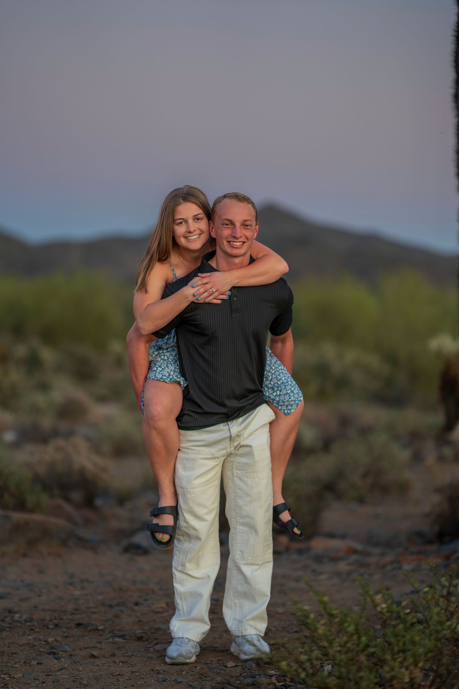 Smiling man giving woman a piggyback ride in Arizona desert at dusk with mountains in background.
