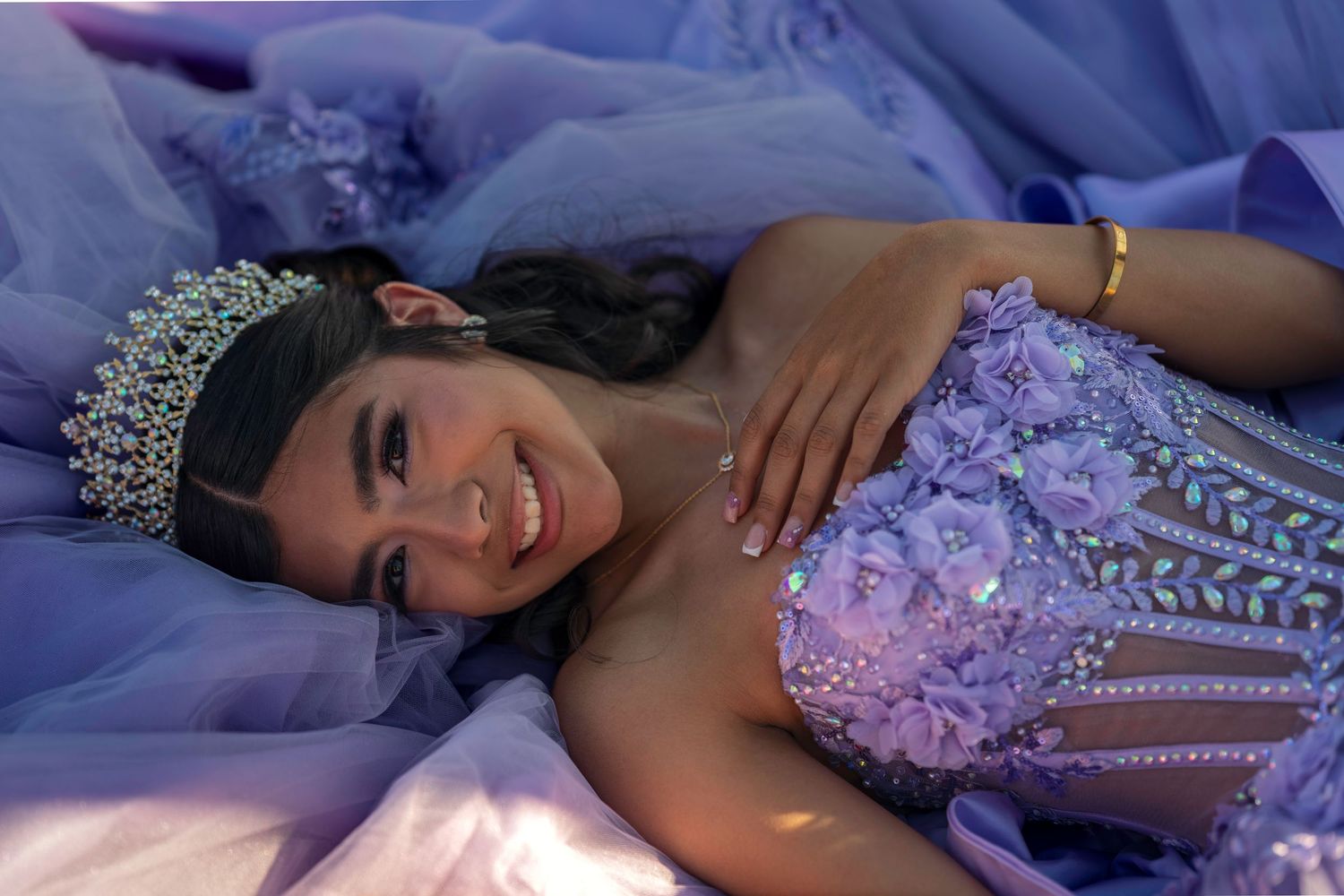 Young woman in lavender quinceañera gown and crystal tiara poses gracefully among purple tulle fabric.