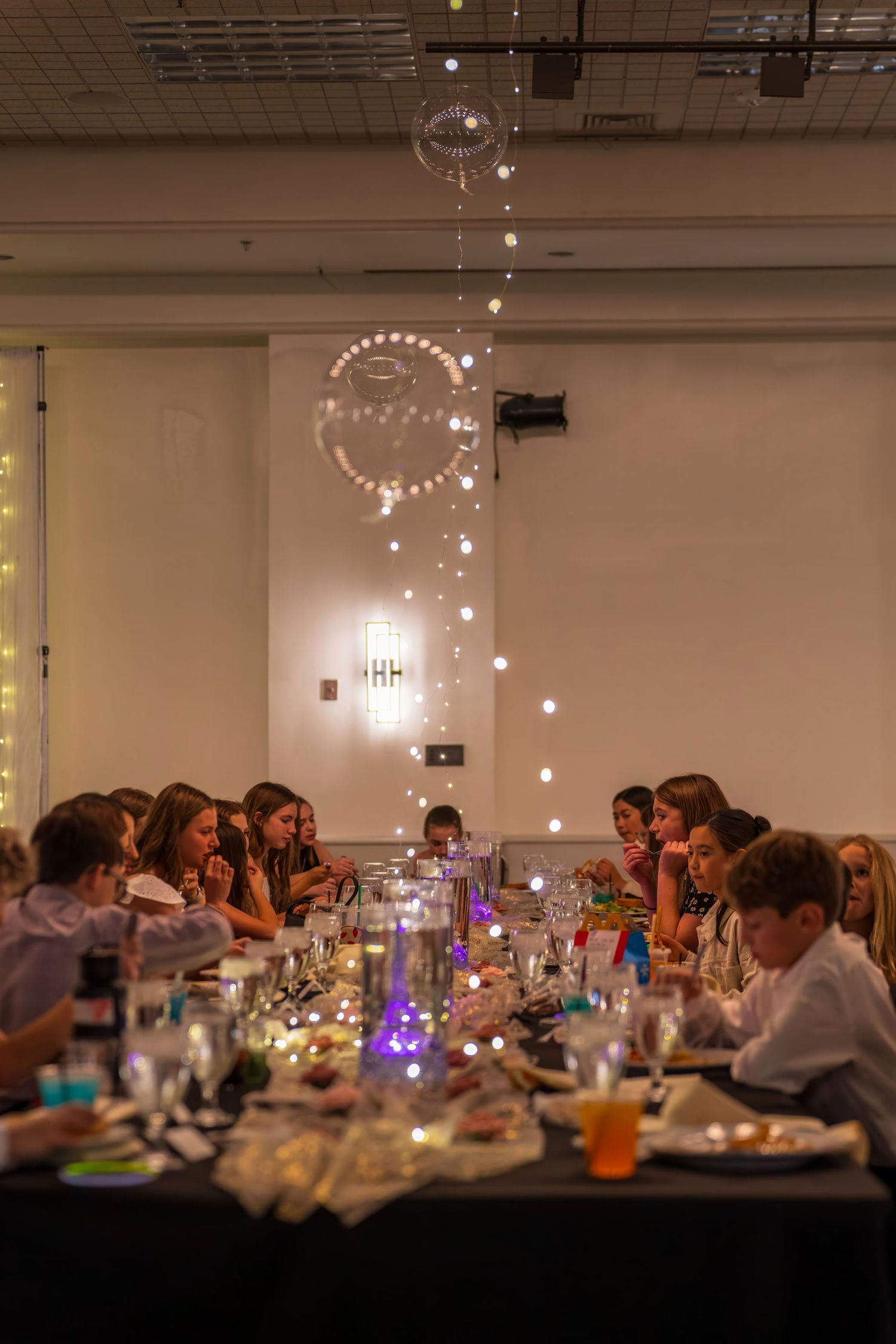 Children and guests dining at a festive banquet table adorned with candles and string lights in an elegant event hall.