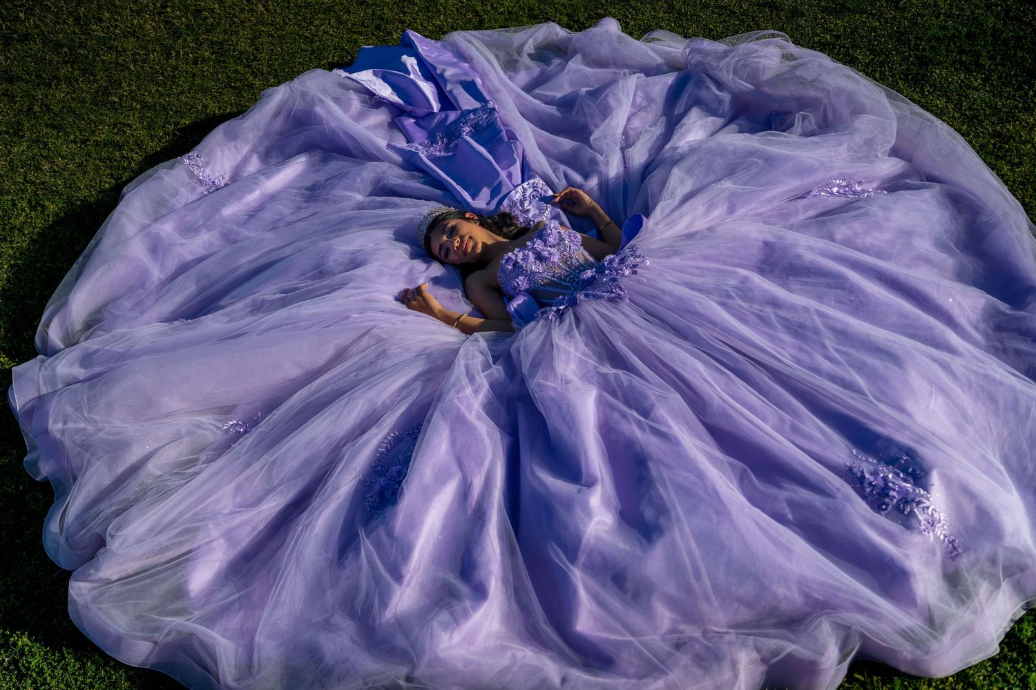 Aerial view of girl in purple quinceañera gown spread across green grass creating floral shape.