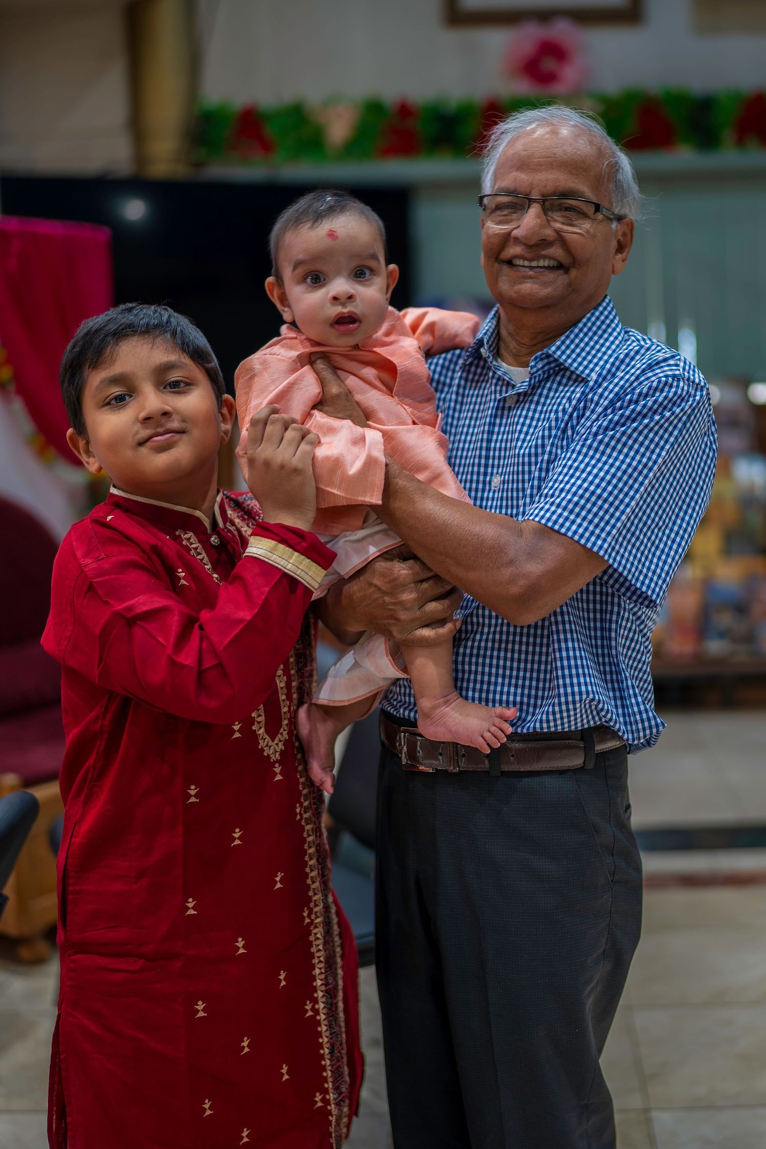 Smiling grandfather holding baby with young boy in red kurta at Indian celebration.