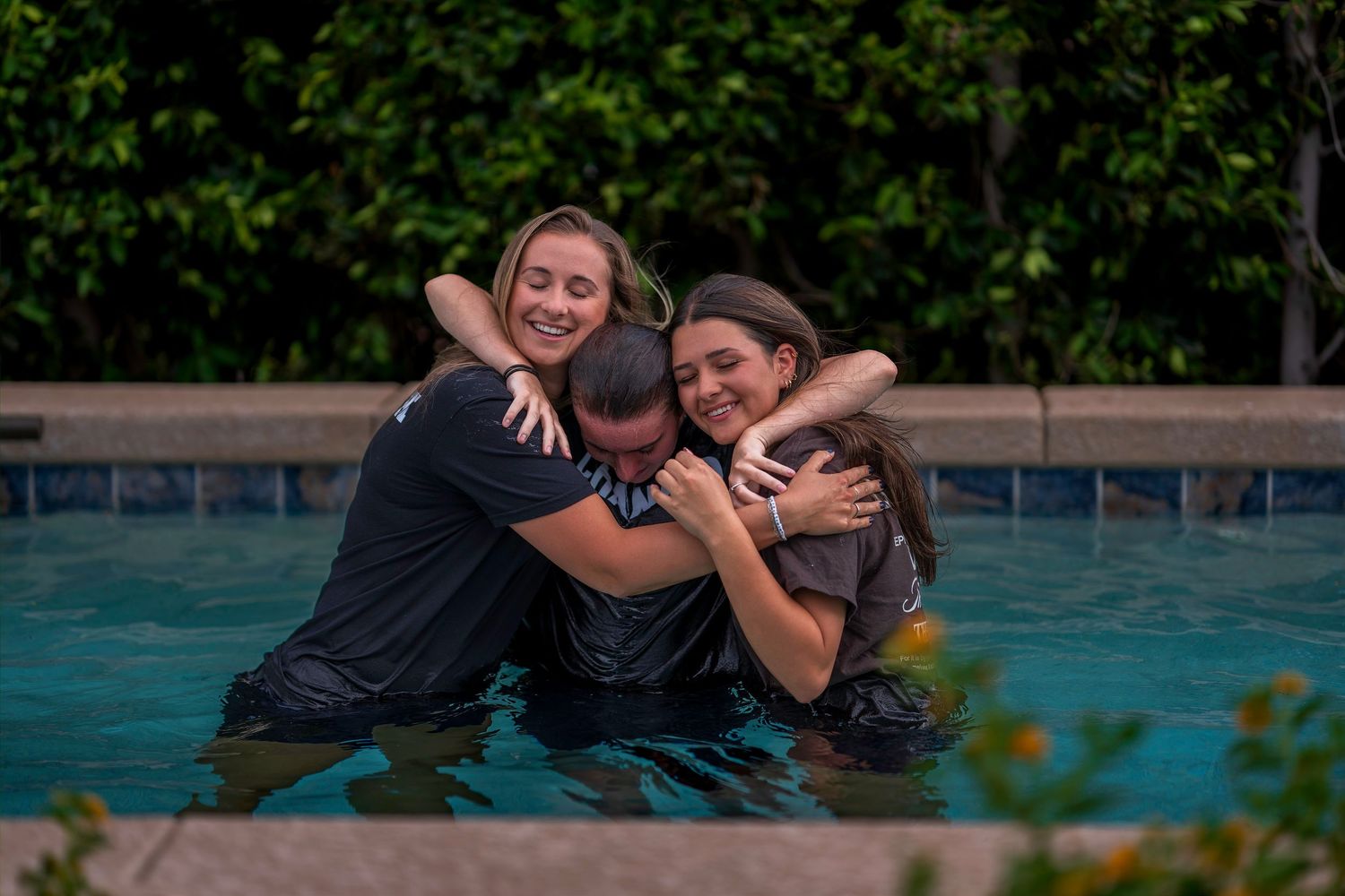 Three people share a joyful group hug while standing in an outdoor swimming pool surrounded by lush greenery.