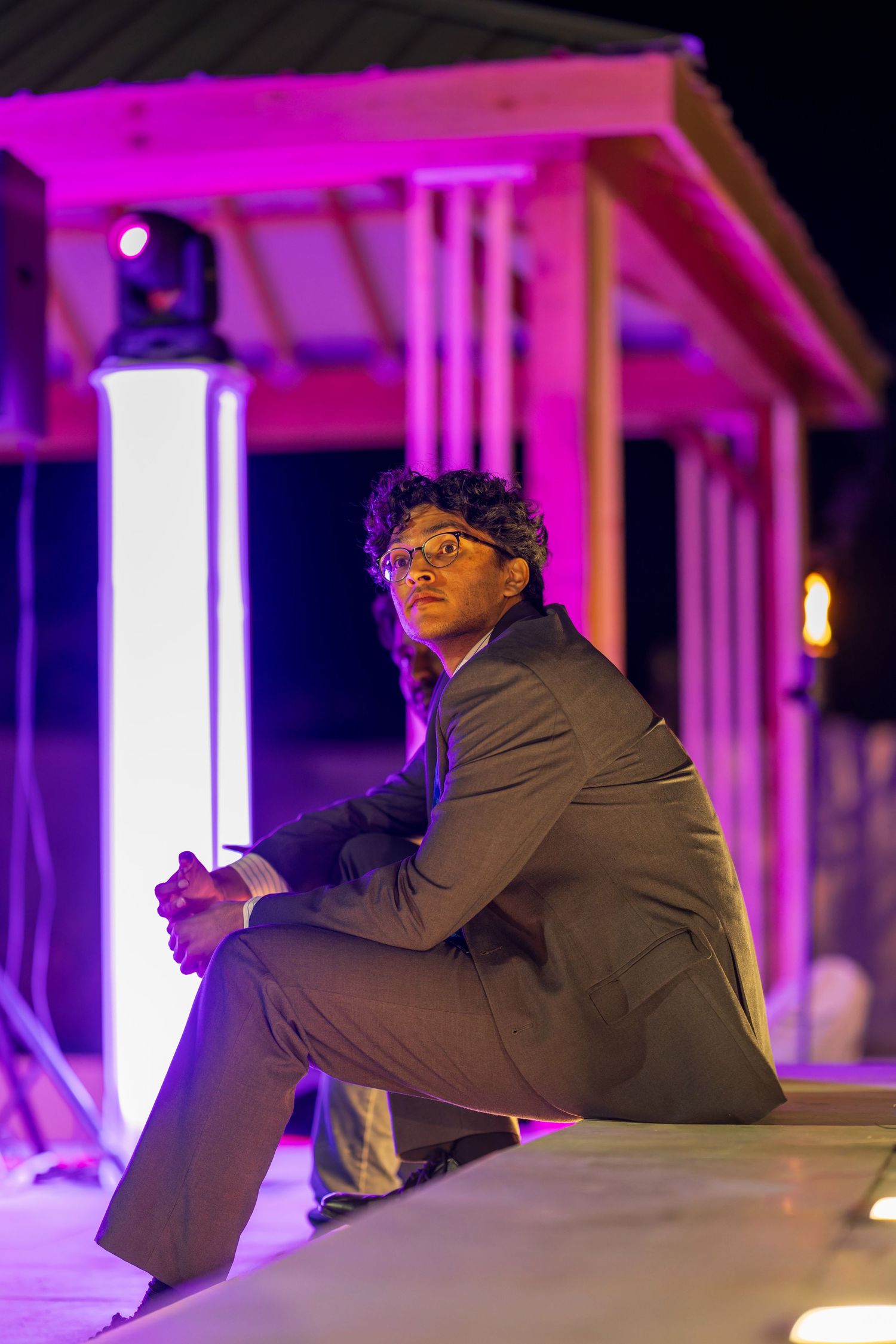 A person in formal attire sits on a ledge under dramatic purple lighting.
