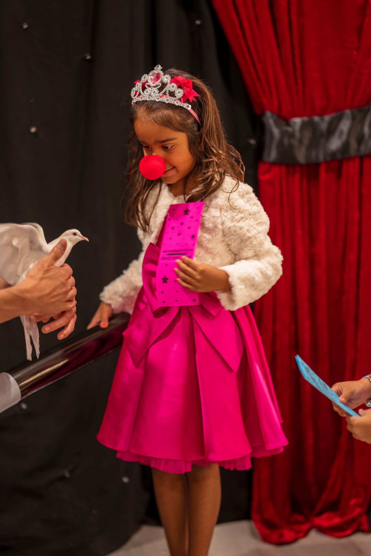 Young performer in a bright pink dress and tiara interacts with a white dove on stage.
