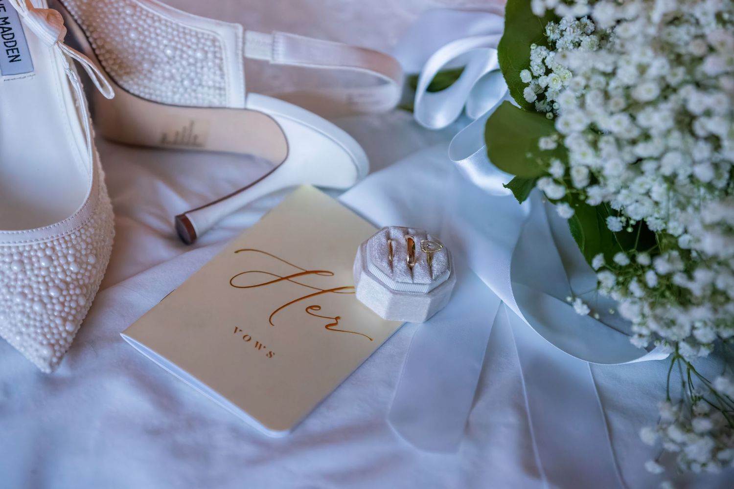 Wedding details featuring elegant bridal shoes, rings, and delicate white flowers on white fabric.