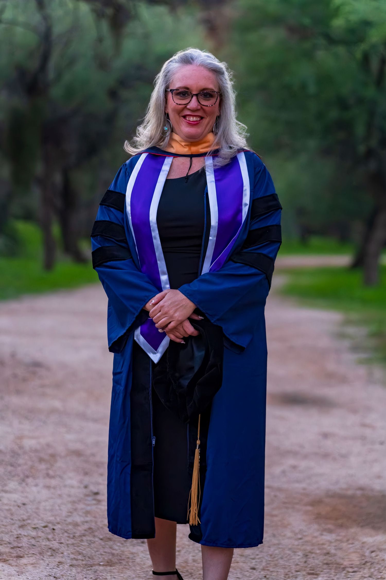 Graduate in traditional academic regalia poses on dirt path surrounded by greenery.