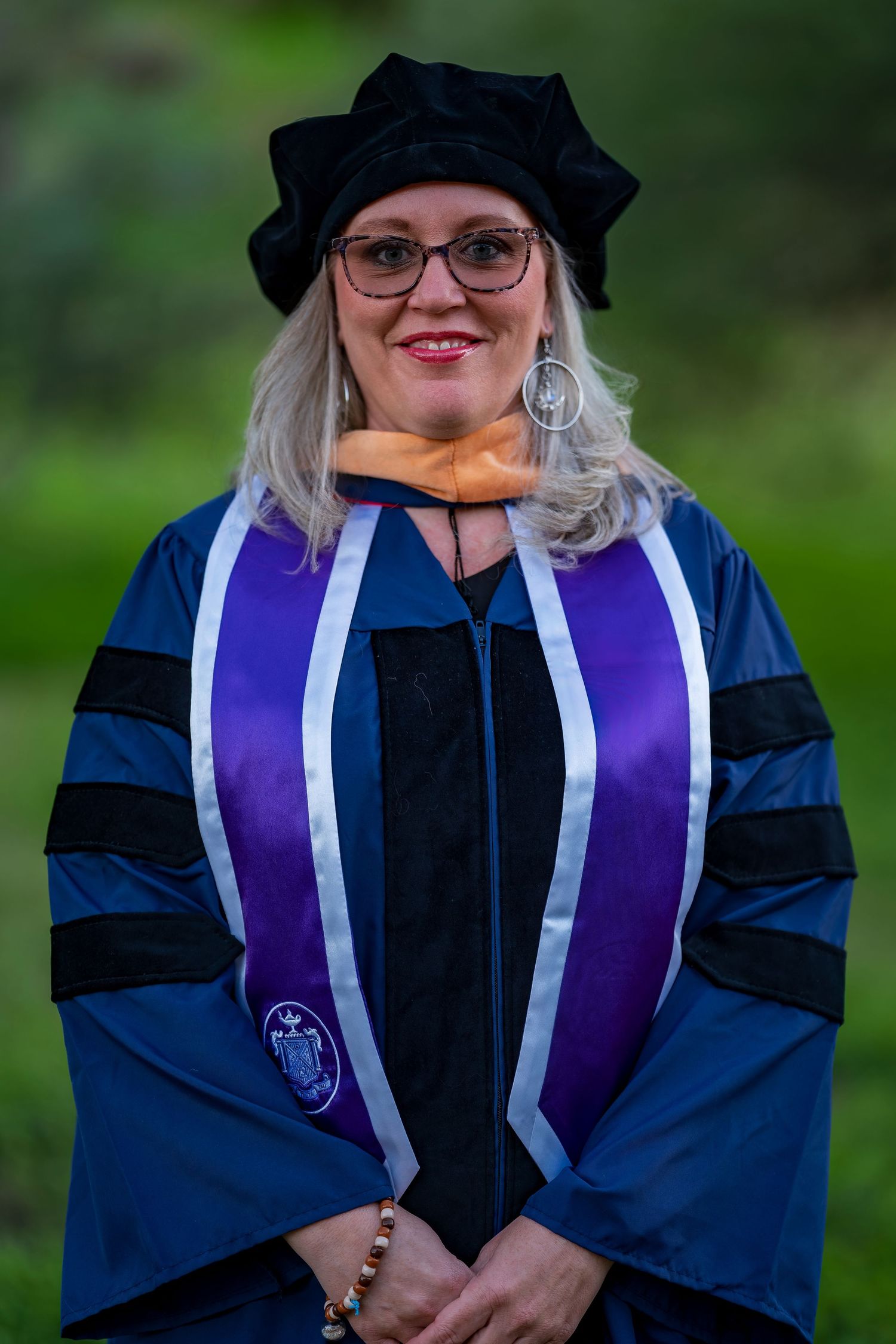 Graduate in blue doctoral regalia and black cap smiles while wearing purple graduation stole.