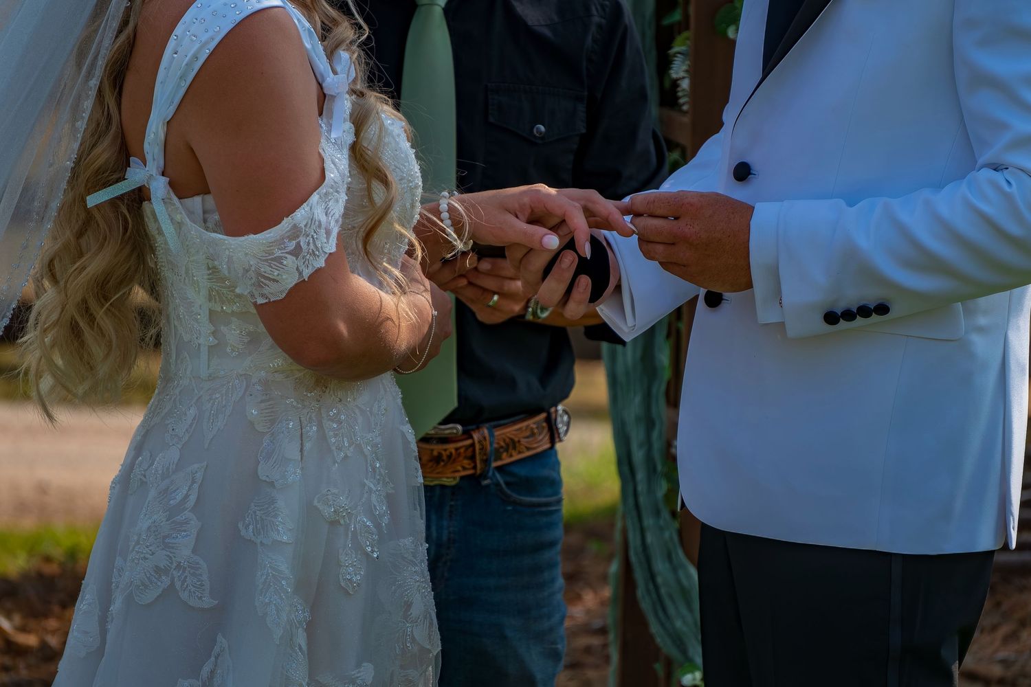Couple exchanging rings during outdoor wedding ceremony under green arbor with floral decorations.