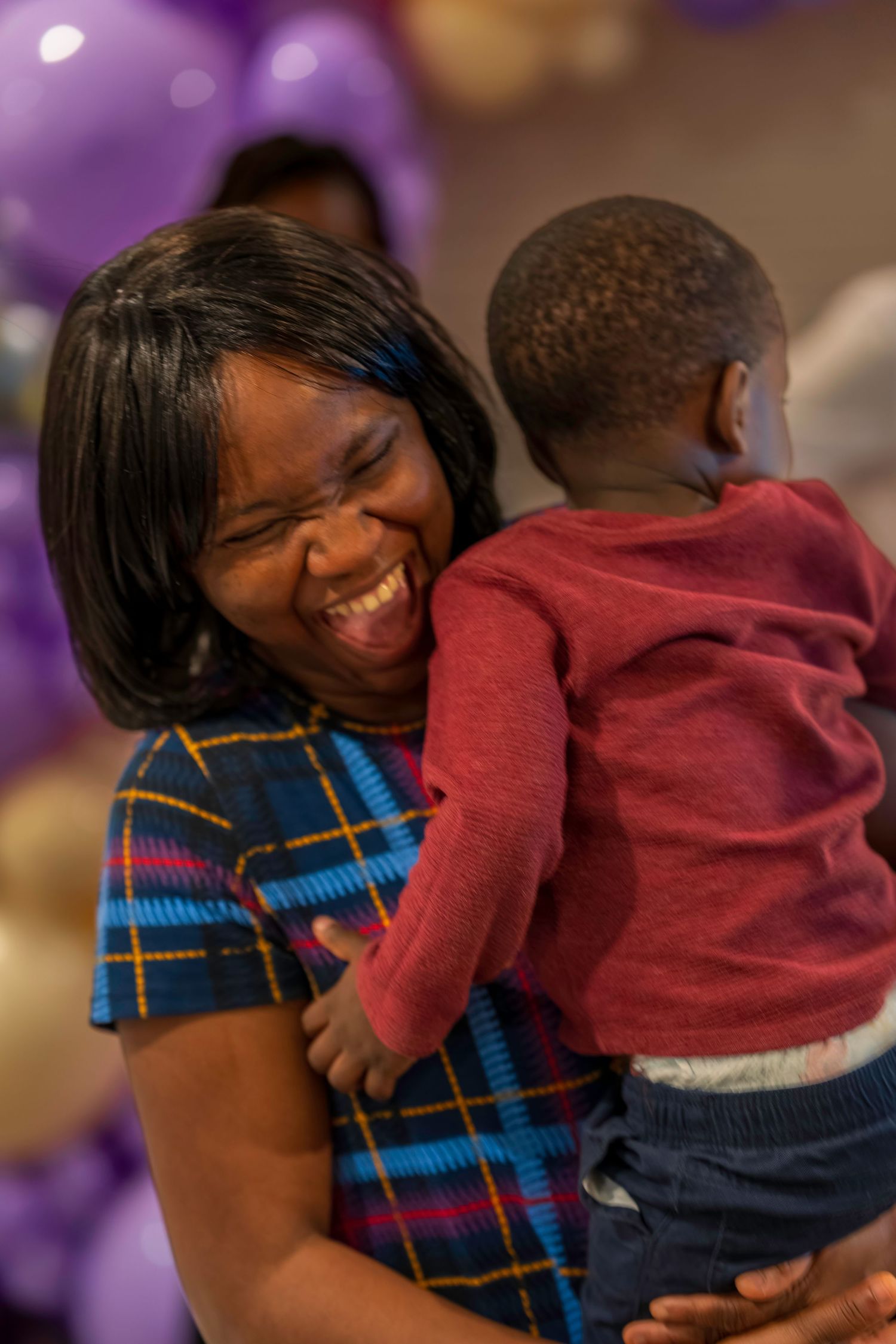 Joyful Black woman in plaid dress laughing while holding a toddler at a festive party with purple and gold balloons.