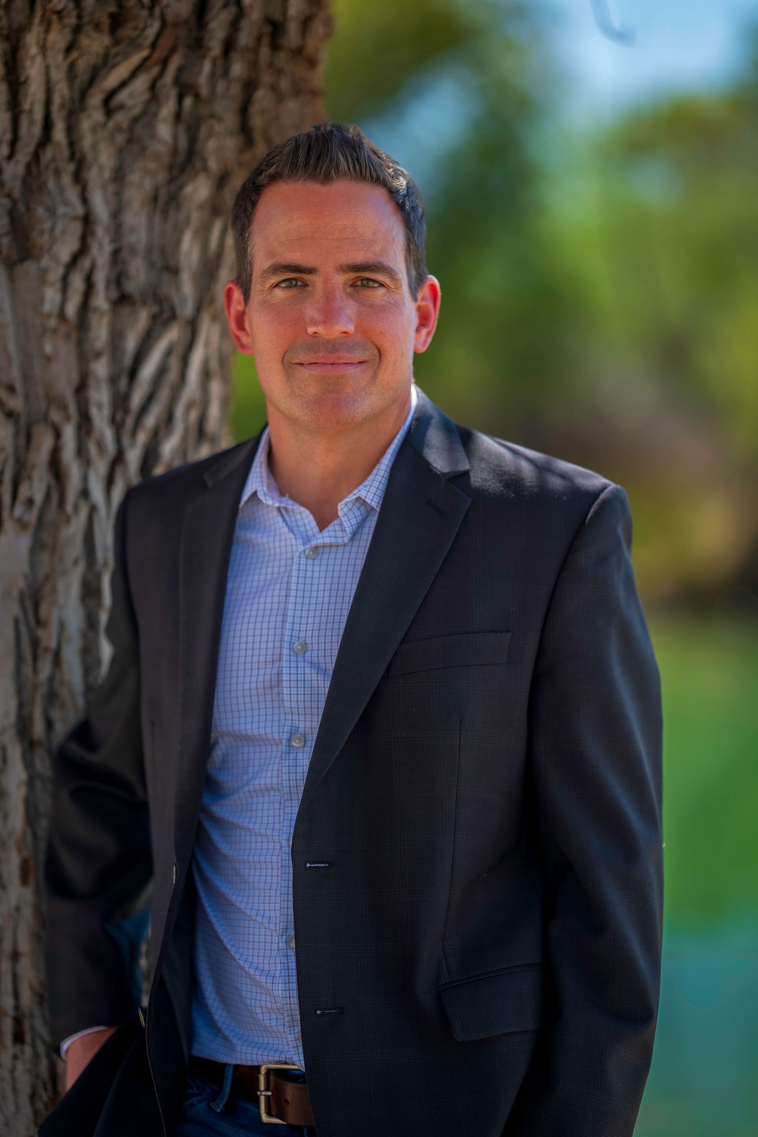 Professional man in dark blazer leaning against tree outdoors with green bokeh background.