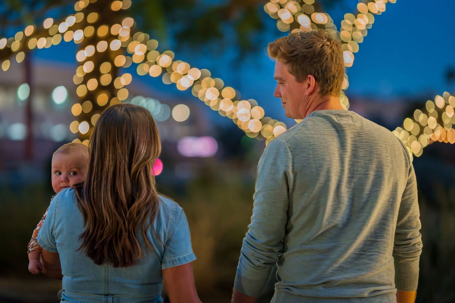 Young family with baby admiring festive bokeh lights outdoors at night.