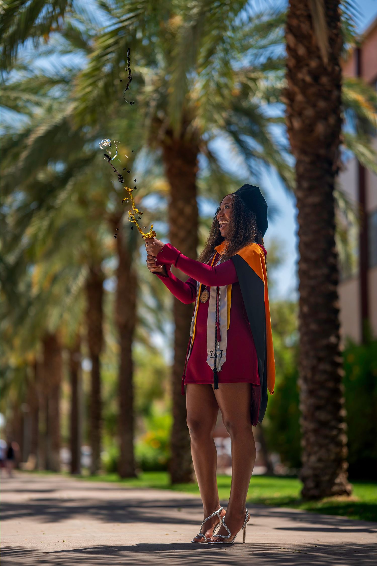Smiling graduate in maroon dress and cap holding flowers among palm trees on a sunny campus pathway.