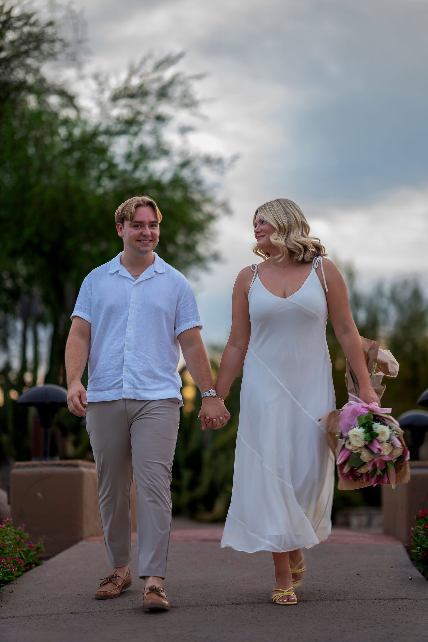 Happy couple in white outfits walking hand-in-hand on a garden path, woman holding a pink and white bouquet.