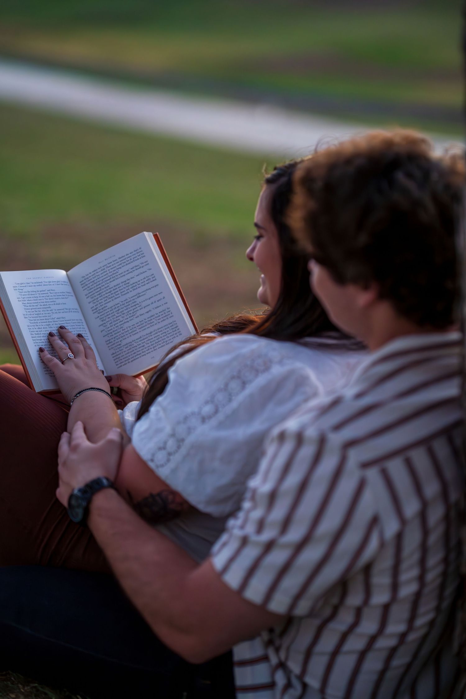 Couple sitting outdoors together reading a book, woman in white lace top holding open pages.
