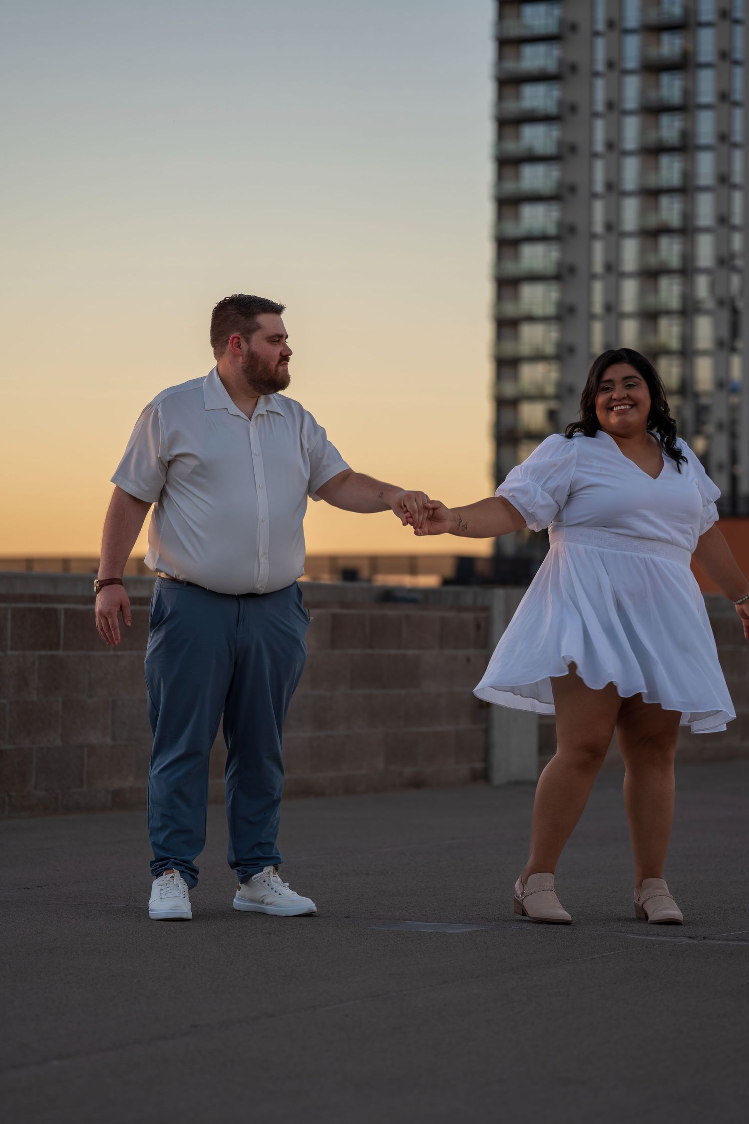 Couple holding hands dancing on rooftop at sunset with city skyline building in background.