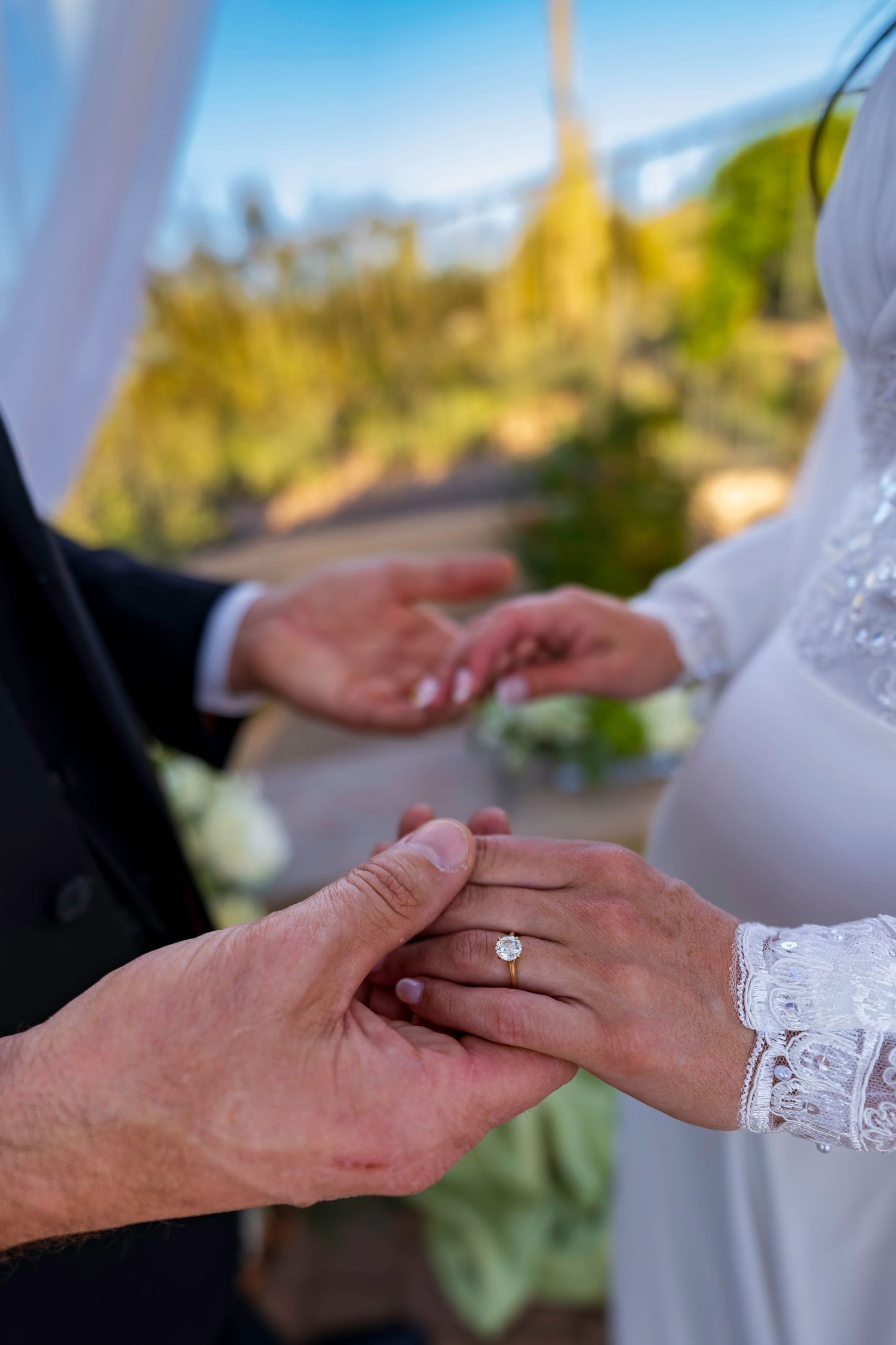 Groom placing diamond engagement ring on bride's finger during outdoor wedding ceremony exchange of vows.
