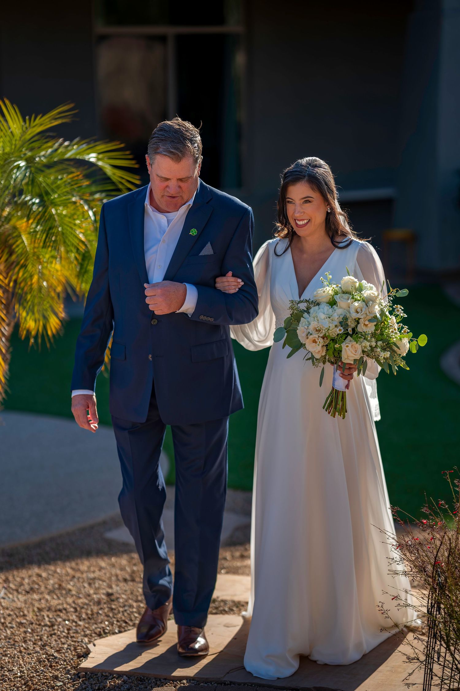 Bride carrying white floral bouquet walks down garden path arm-in-arm with escort in navy blue suit.