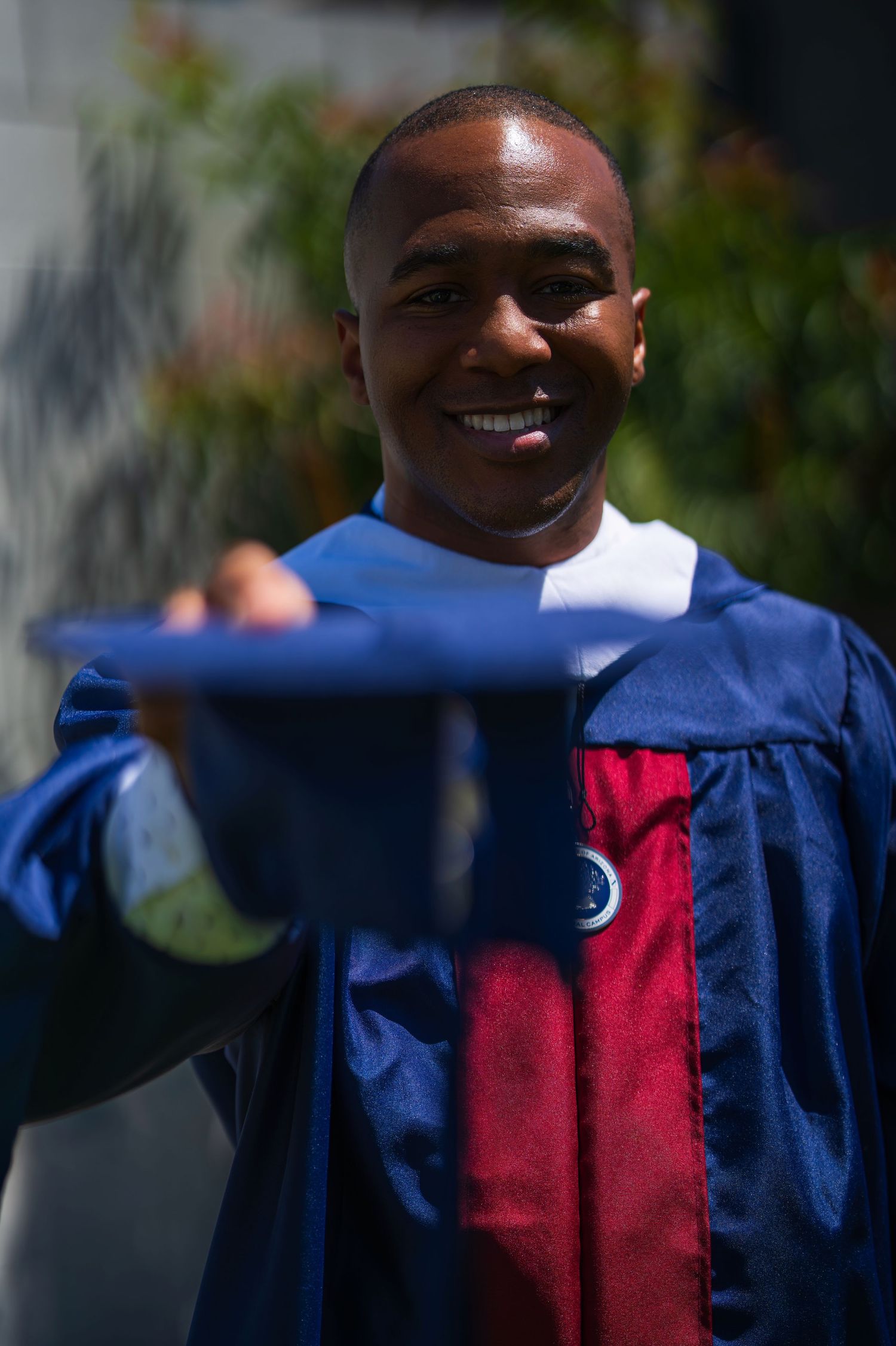 Graduate in blue and red academic regalia smiling on a sunny day.
