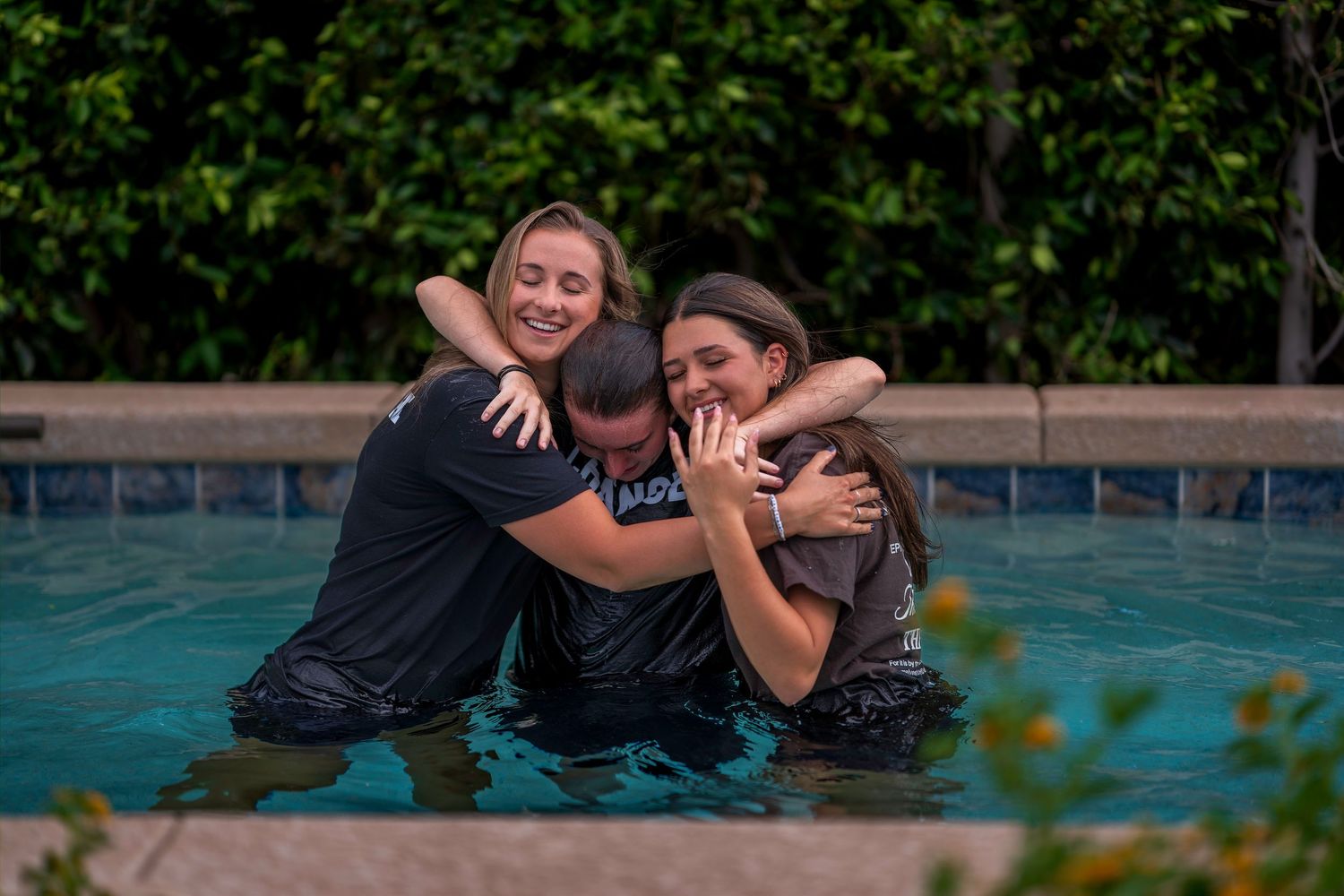 Three people hugging joyfully in a swimming pool, sharing a warm embrace outdoors.