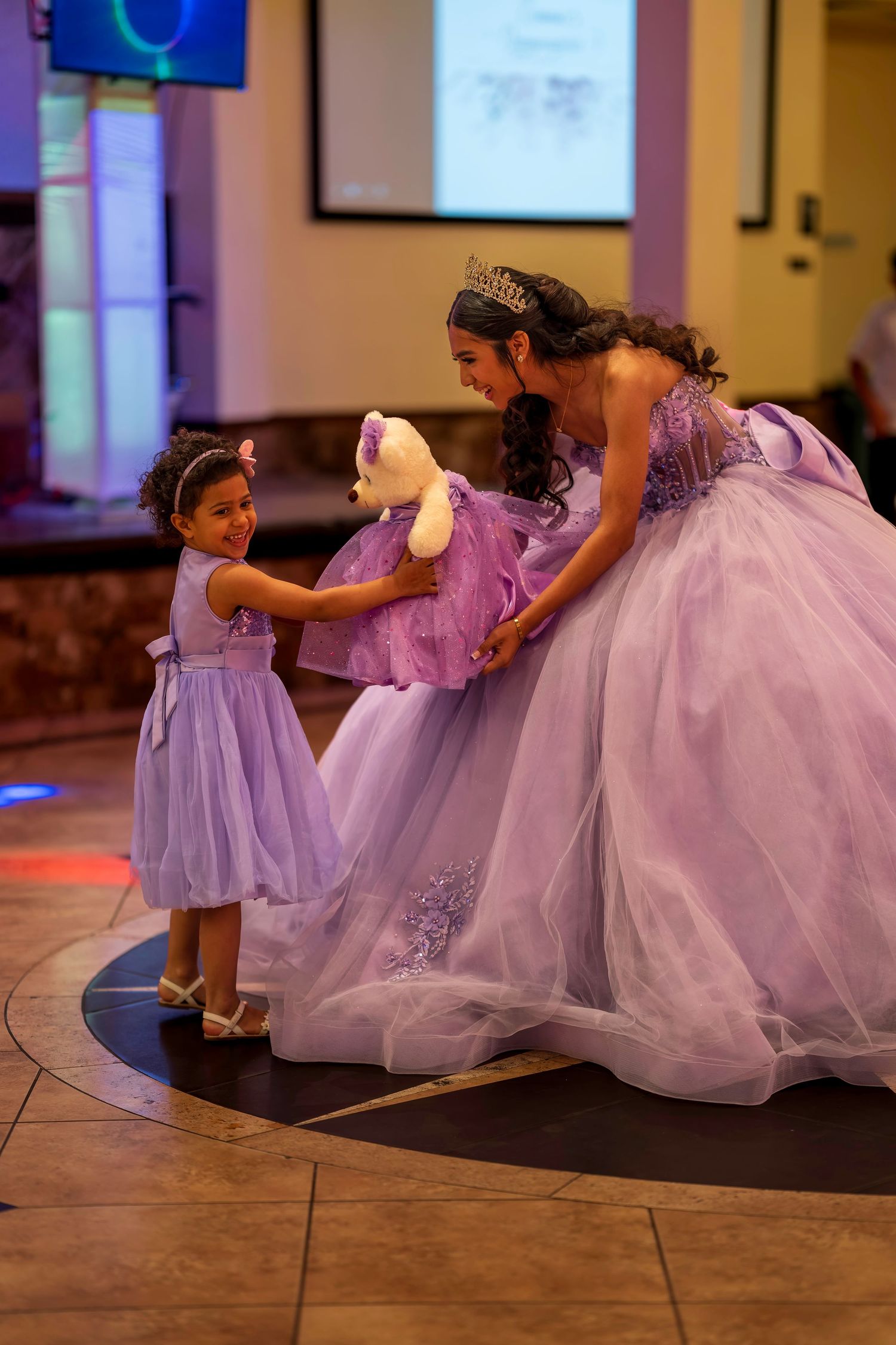 A quinceañera girl in a lavender ball gown shares a sweet moment with a young flower girl on the dance floor.