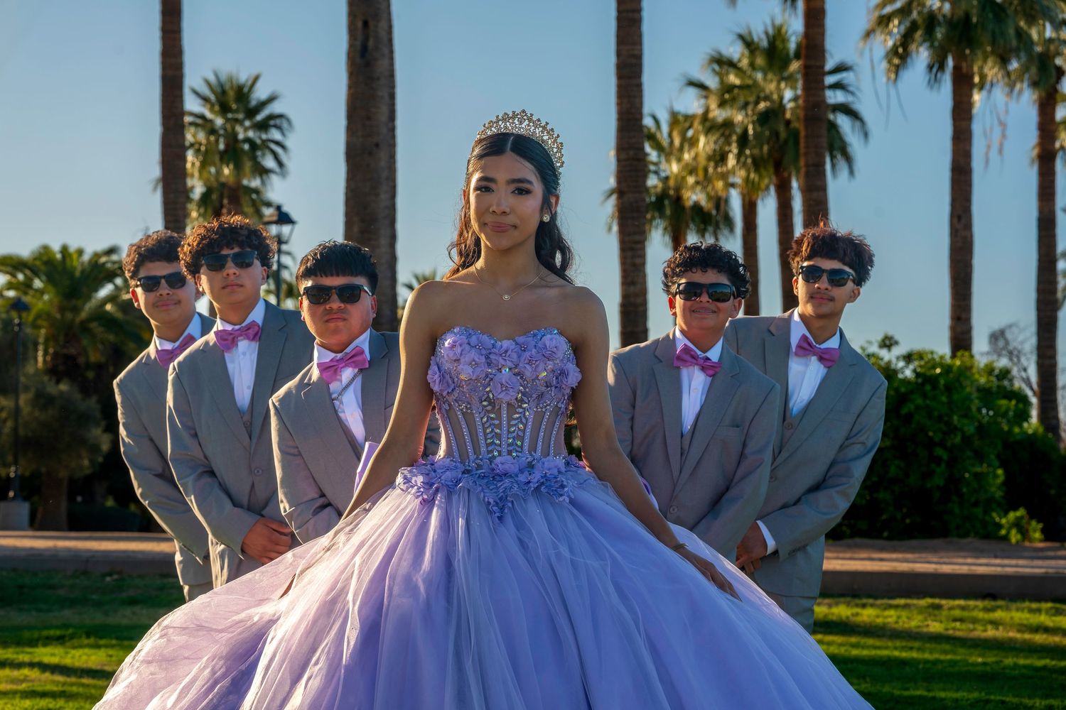 Young woman in lavender quinceañera ball gown and tiara poses with five young men in gray suits and pink bow ties outdoors.