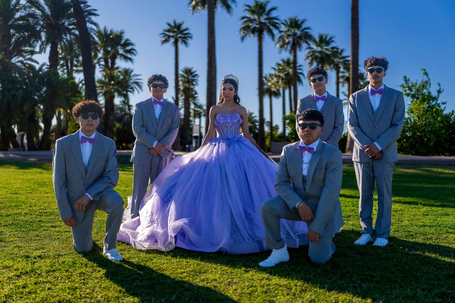 Young woman in lavender quinceañera gown poses with chambelanes in gray suits on green lawn with palm trees.