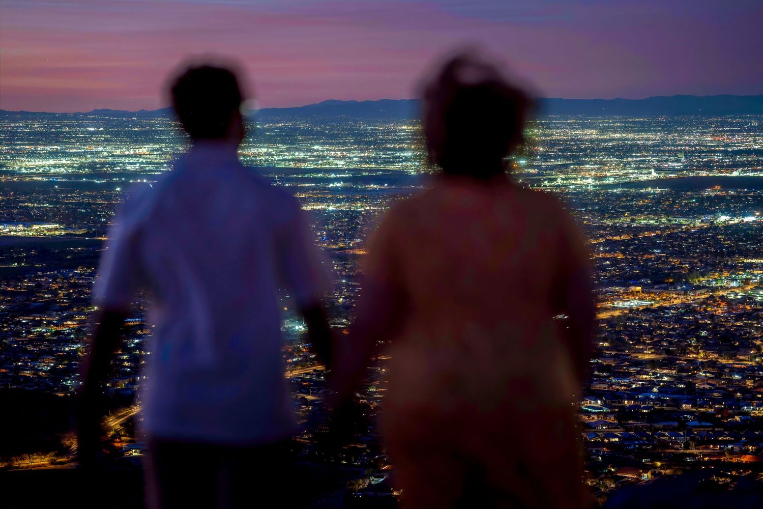 Couple holding hands viewing stunning city lights from hilltop at twilight.