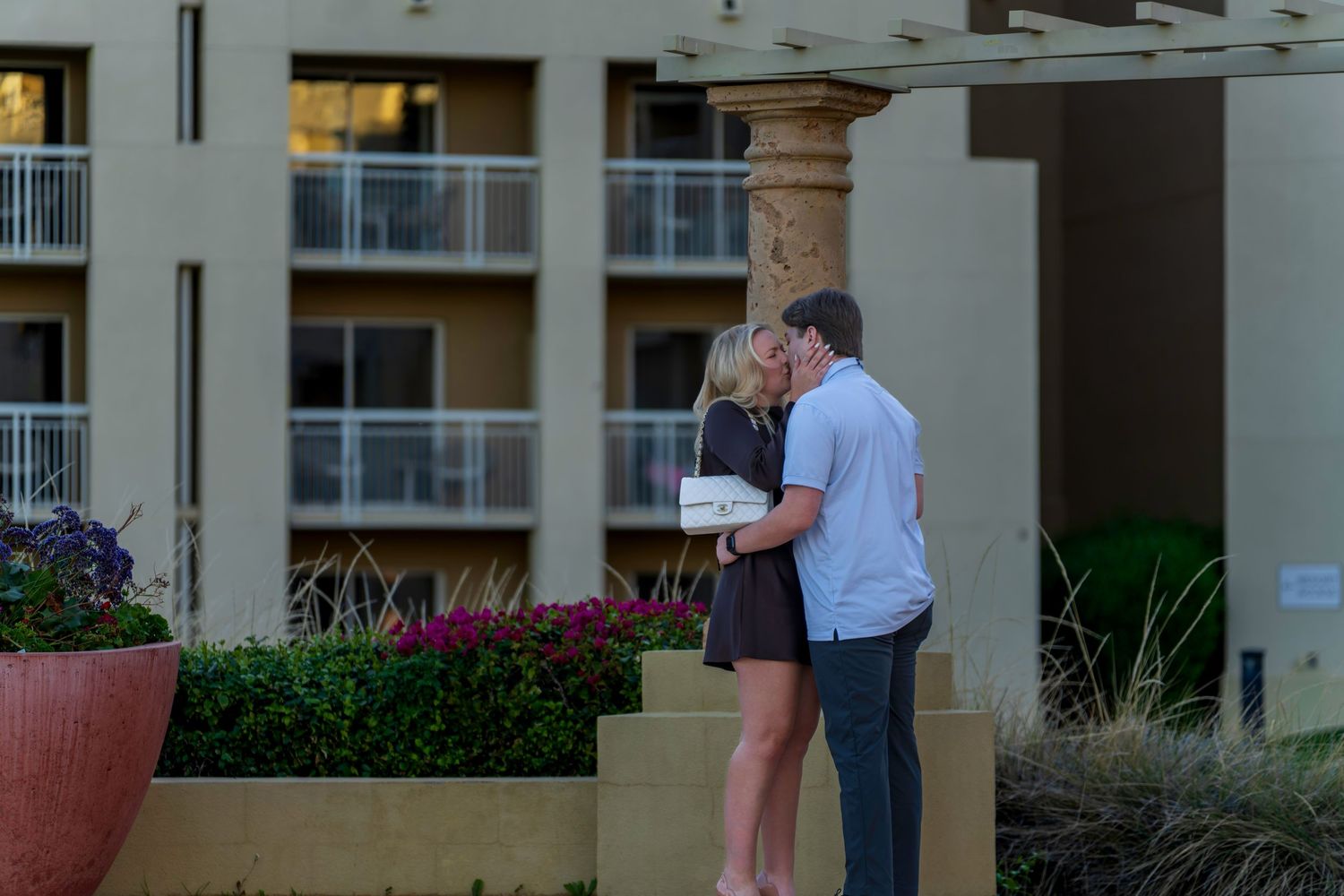 Couple sharing a romantic kiss outside a luxury resort building with balconies and colorful flowers.