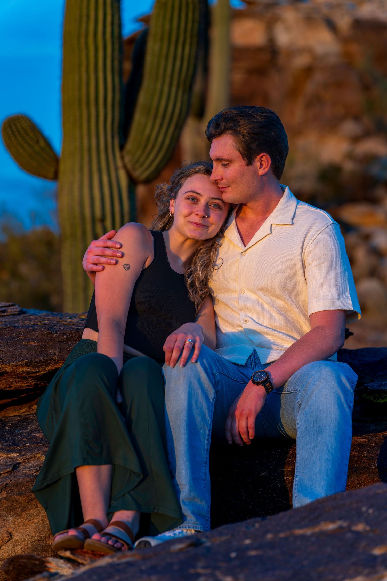 Couple sitting together on desert rocks with a saguaro cactus backdrop at dusk.