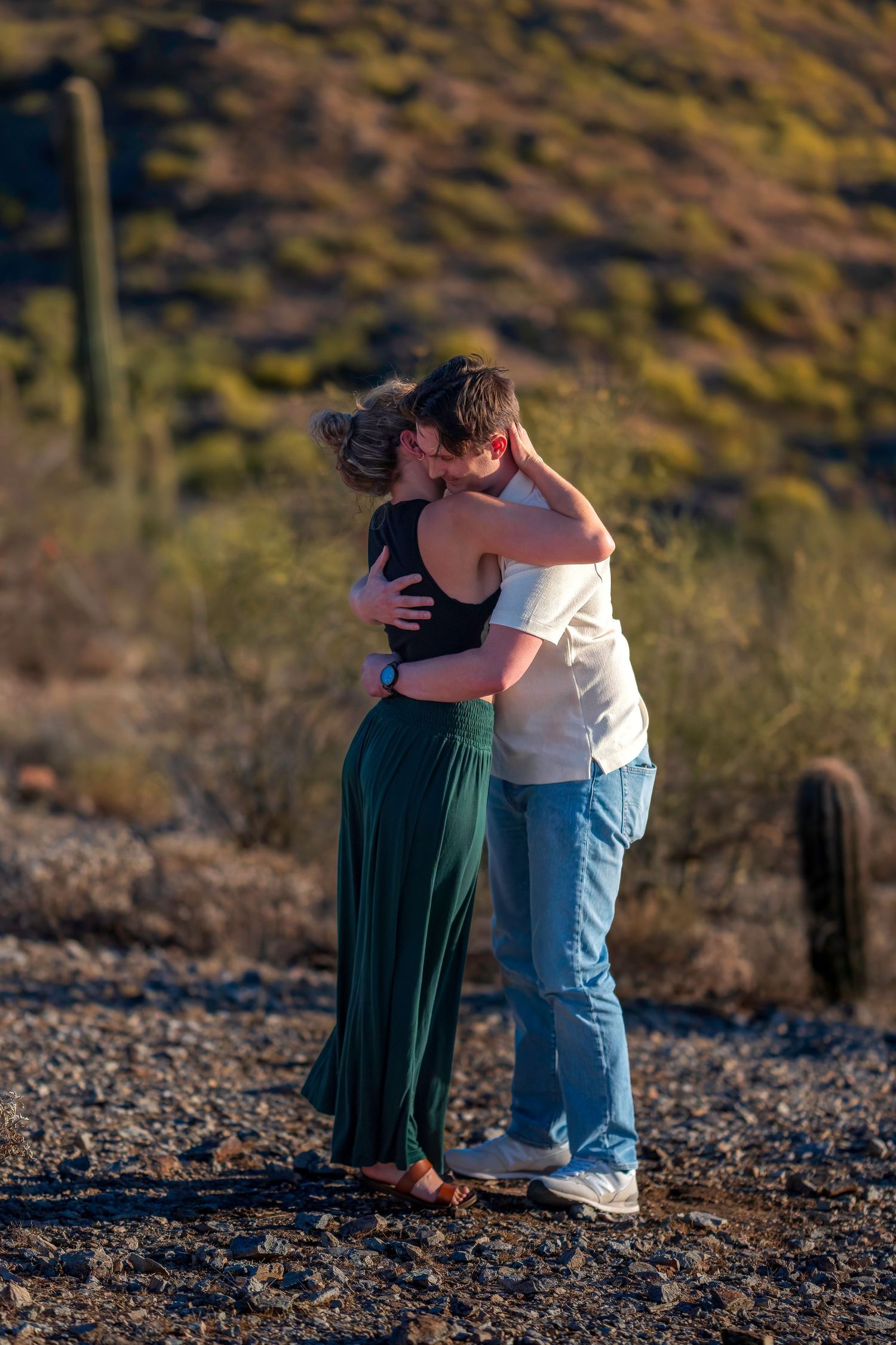 Couple sharing a romantic kiss while embracing in a desert landscape with saguaro cacti at golden hour.