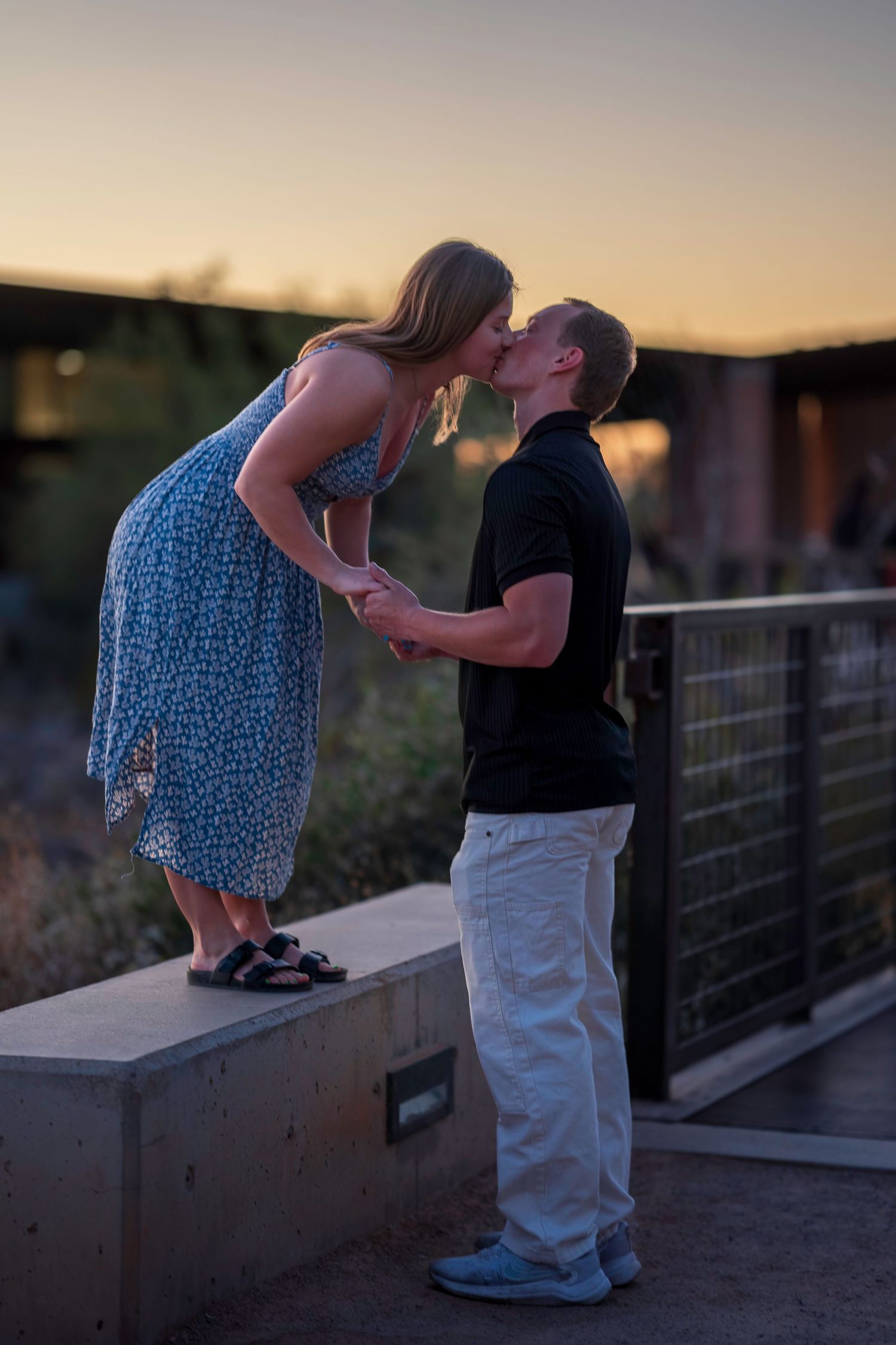 A couple kisses at sunset, she stands on a ledge in a blue floral dress while he holds her hands.