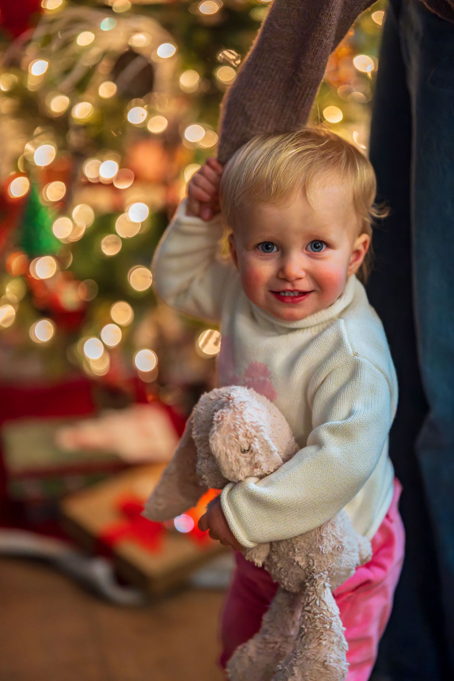 Baby in white sweater holding teddy bear with Christmas lights in background.