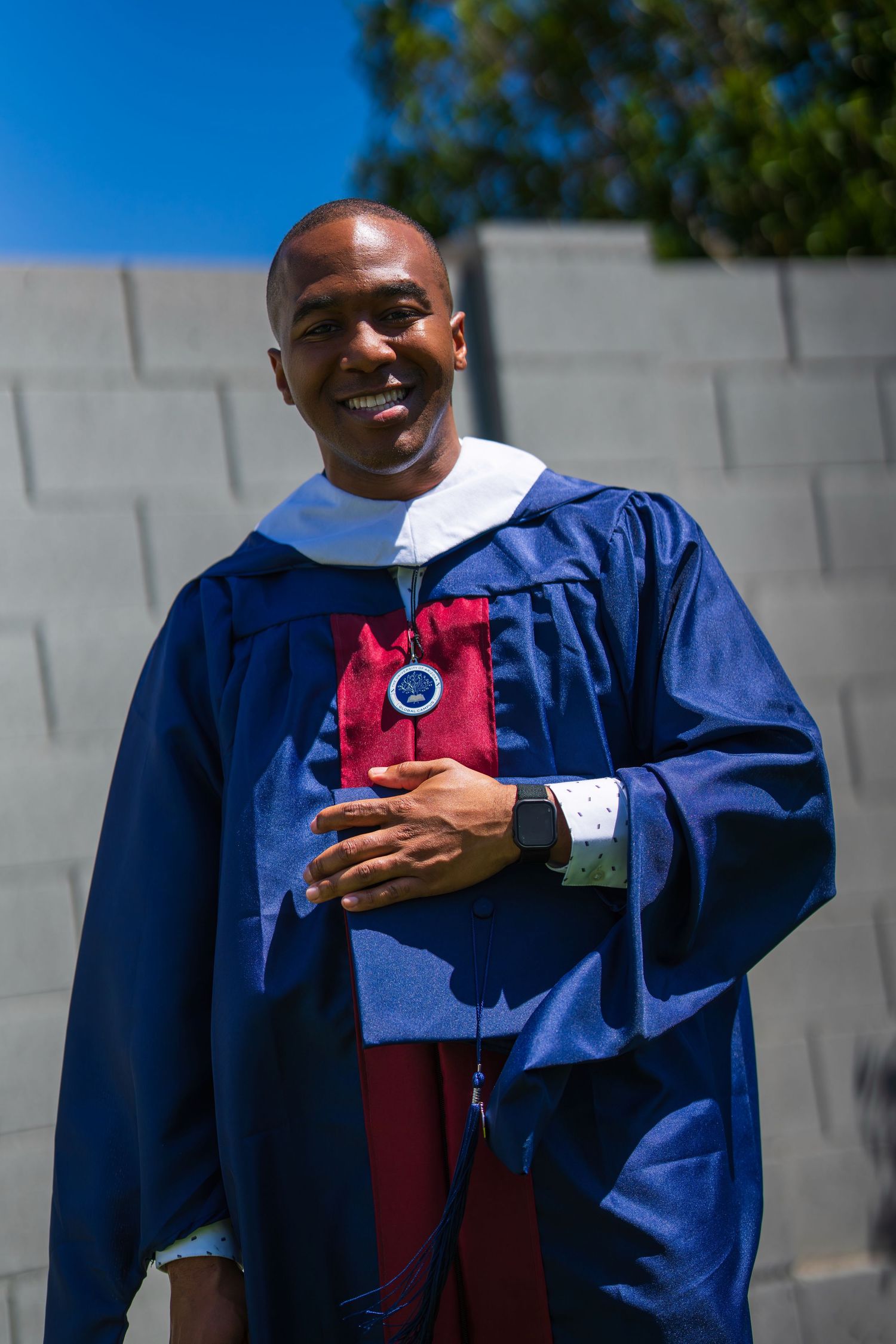 Graduate wearing blue and red academic regalia smiling outdoors on a sunny day.