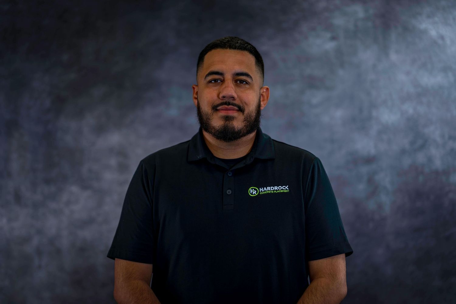 Professional headshot of person in dark polo with company logo against gray background.