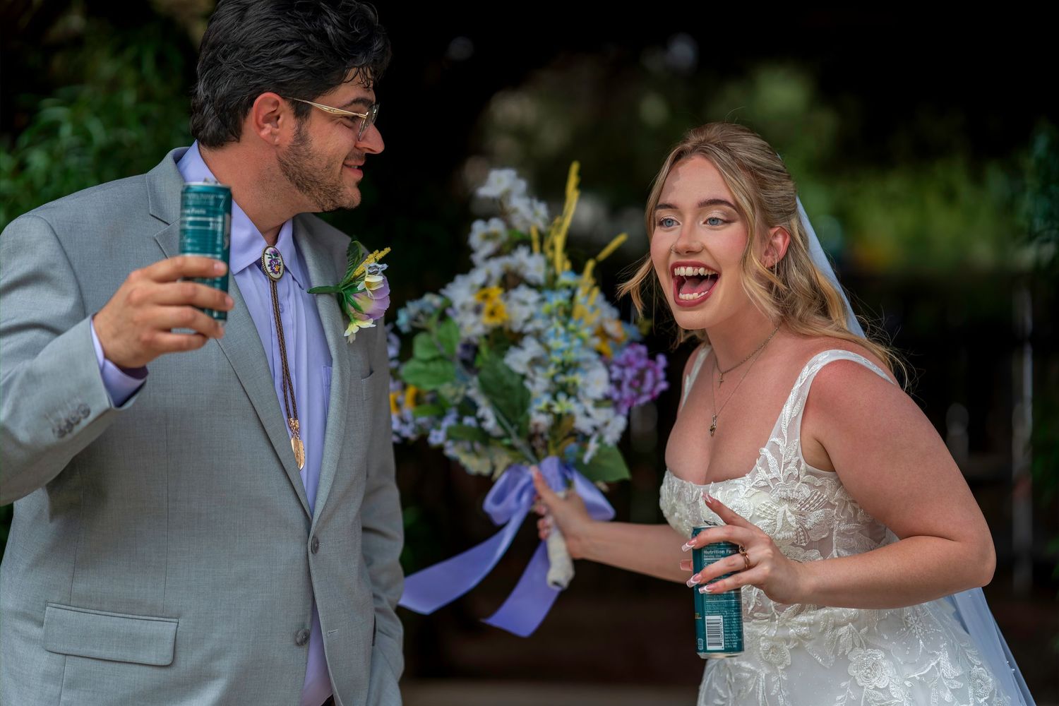 Joyful bride and groom laughing together holding drinks at outdoor wedding ceremony.