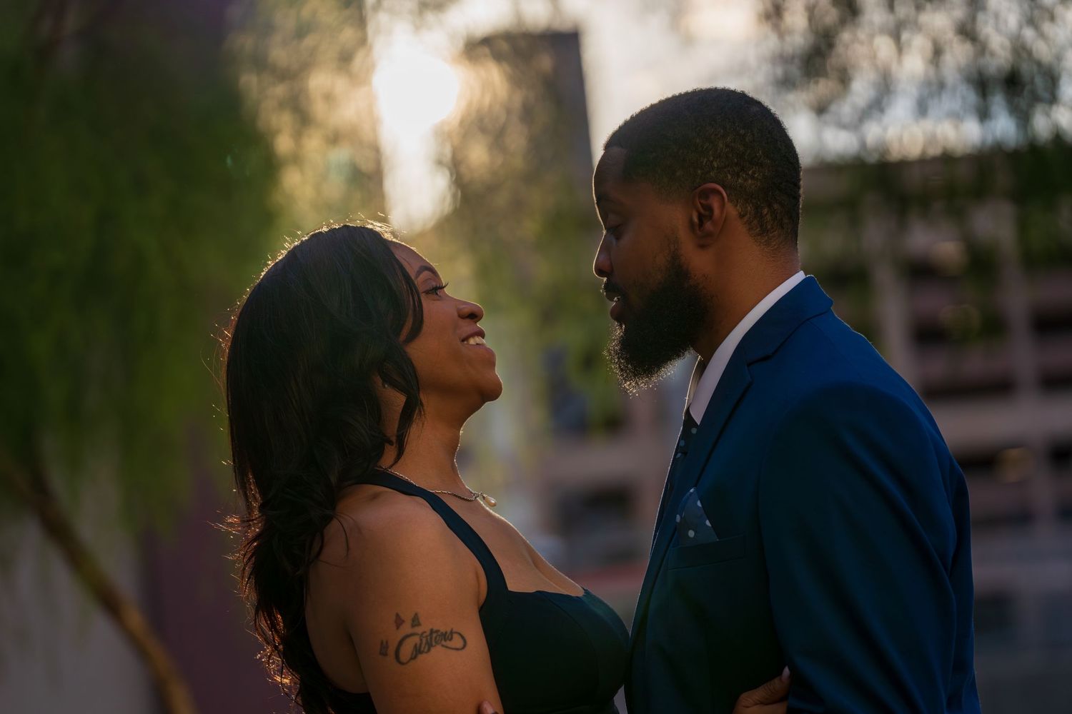 A couple shares a romantic moment outdoors at sunset, with the woman smiling up at her partner in an urban setting.