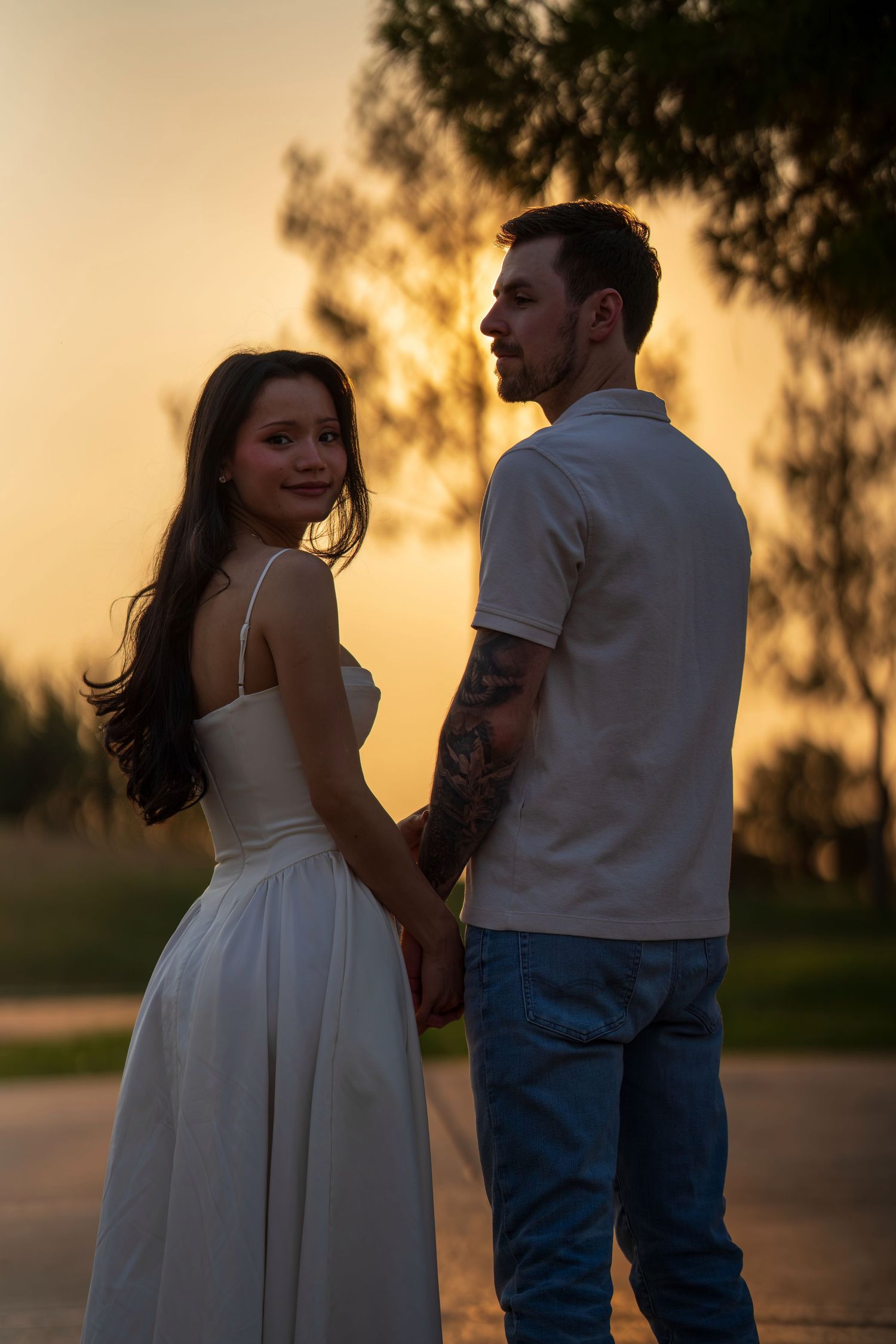 Couple holding hands at sunset, bride in white dress looks back while groom has tattoo sleeve.