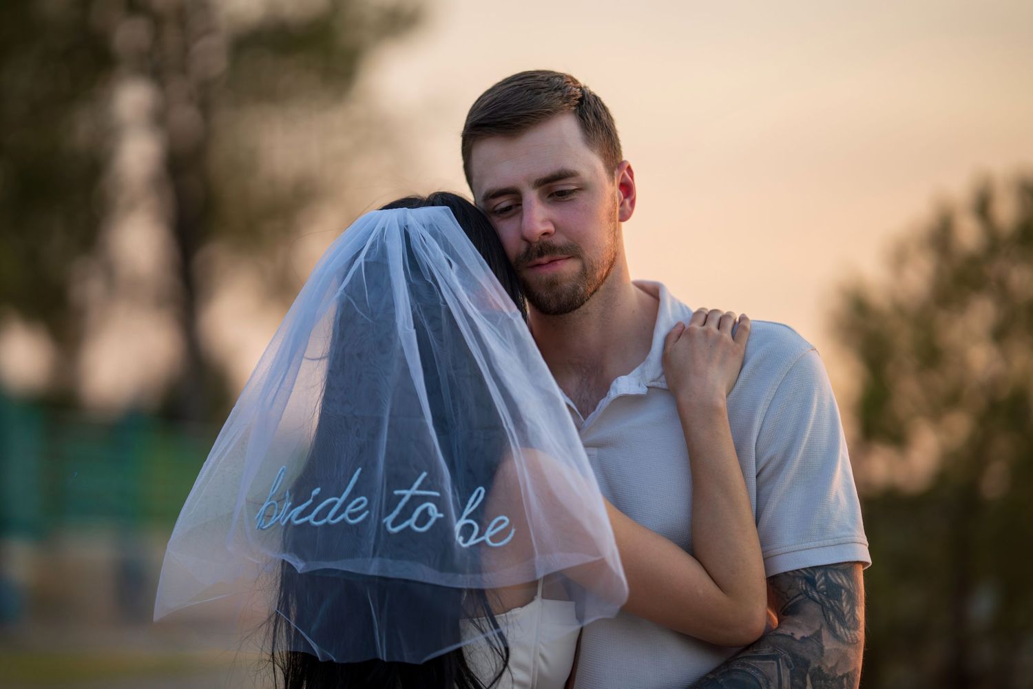 Man embracing woman wearing a bride-to-be veil at sunset, both sharing a tender moment.