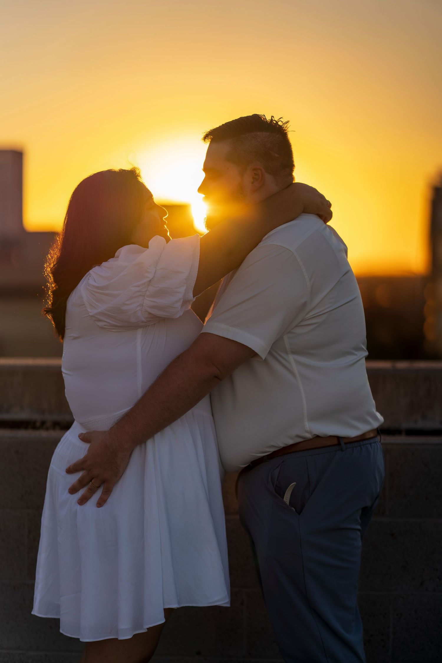 Couple embracing romantically at golden sunset, woman in white dress and man in white shirt, silhouetted against orange sky.