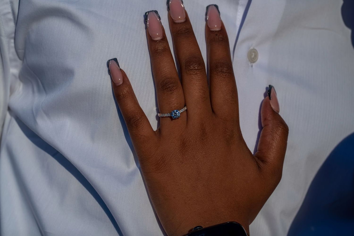 Close-up of engagement ring with blue stone on woman's hand with French tip nails against white shirt.