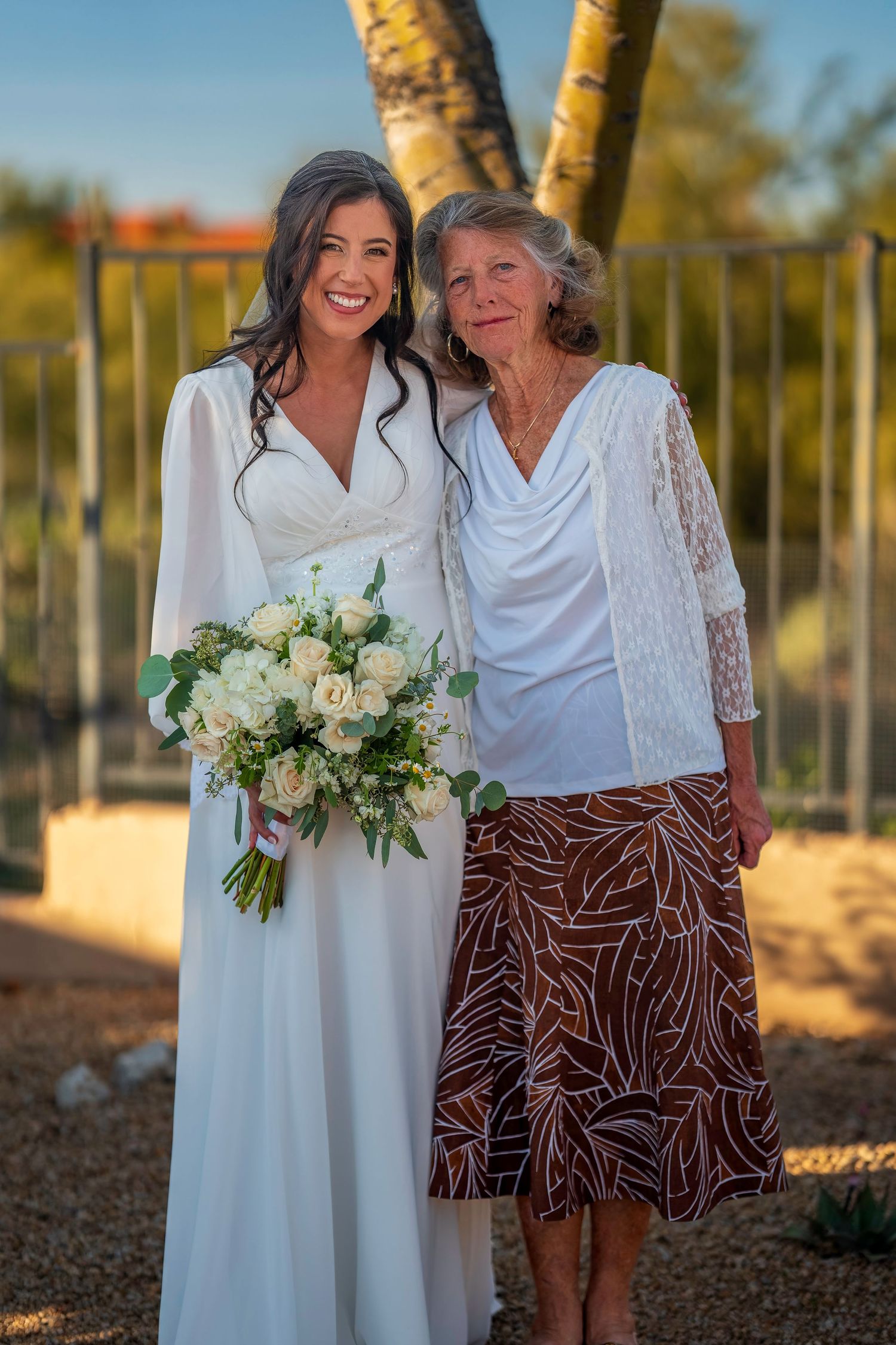 Smiling bride in white gown holding roses posing with older woman at outdoor wedding.