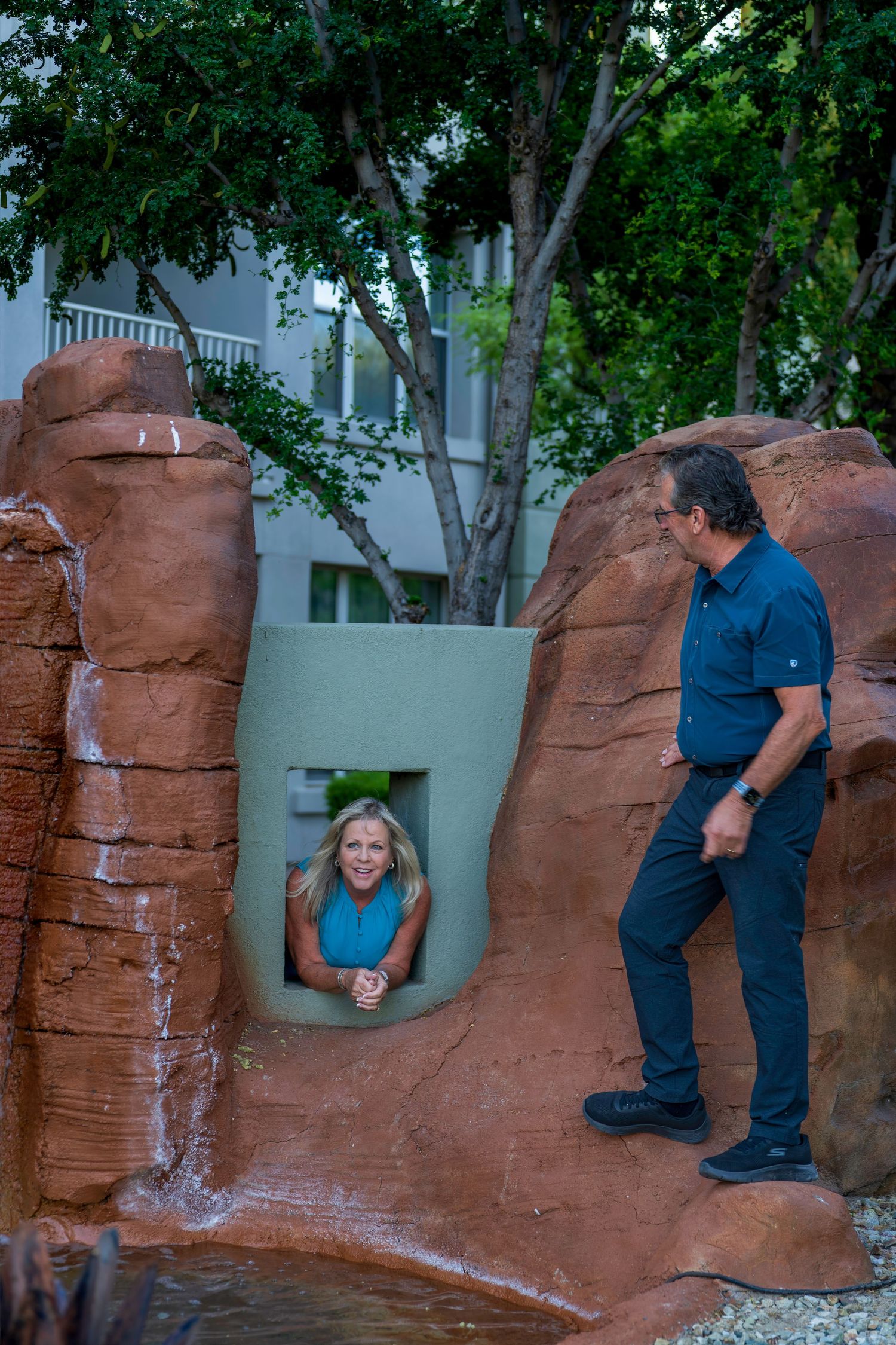 A woman peeks through a decorative rock tunnel while a man stands nearby at a resort.