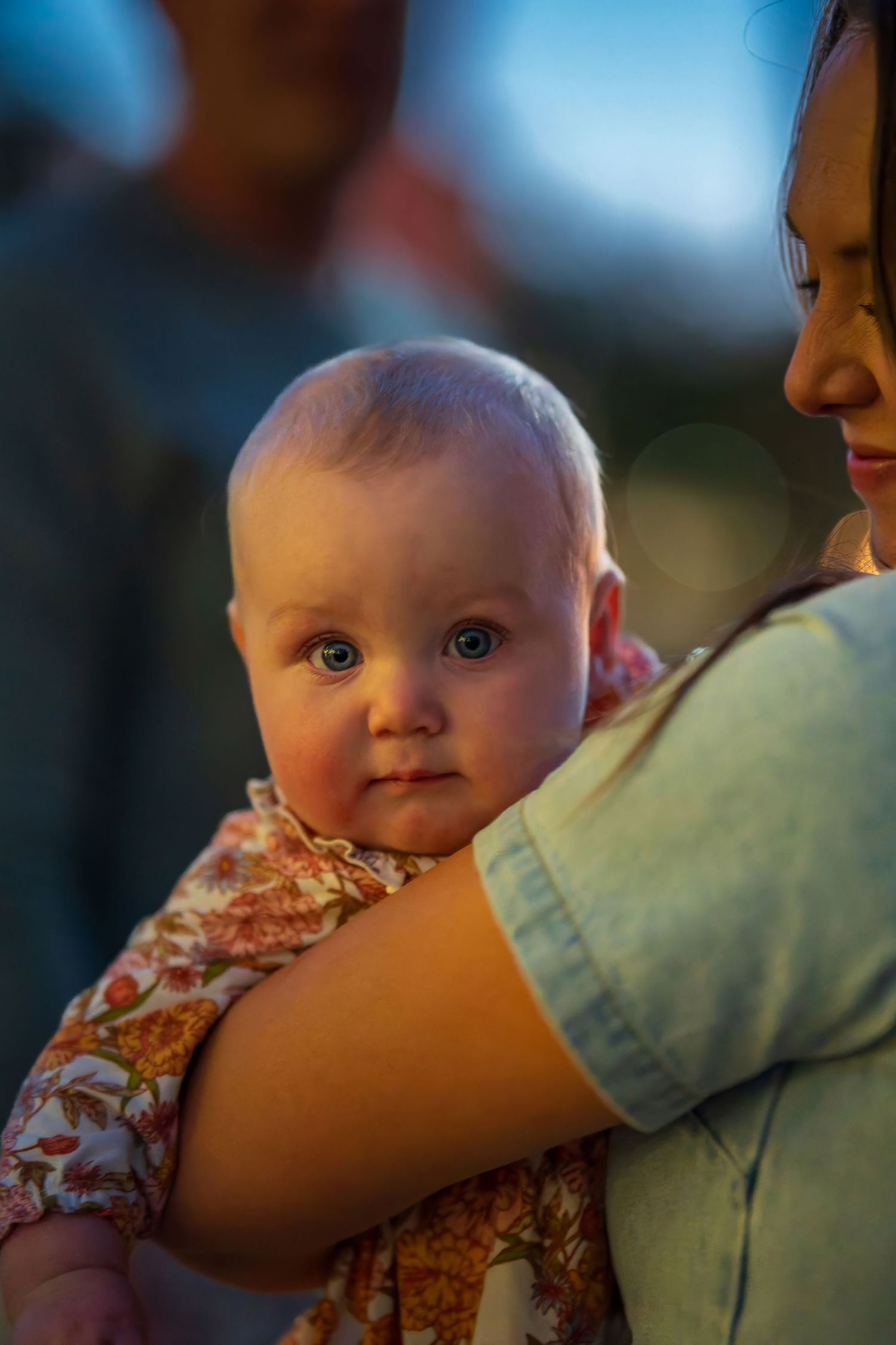 A wide-eyed baby in a floral outfit is held by a woman in a light blue shirt at dusk.