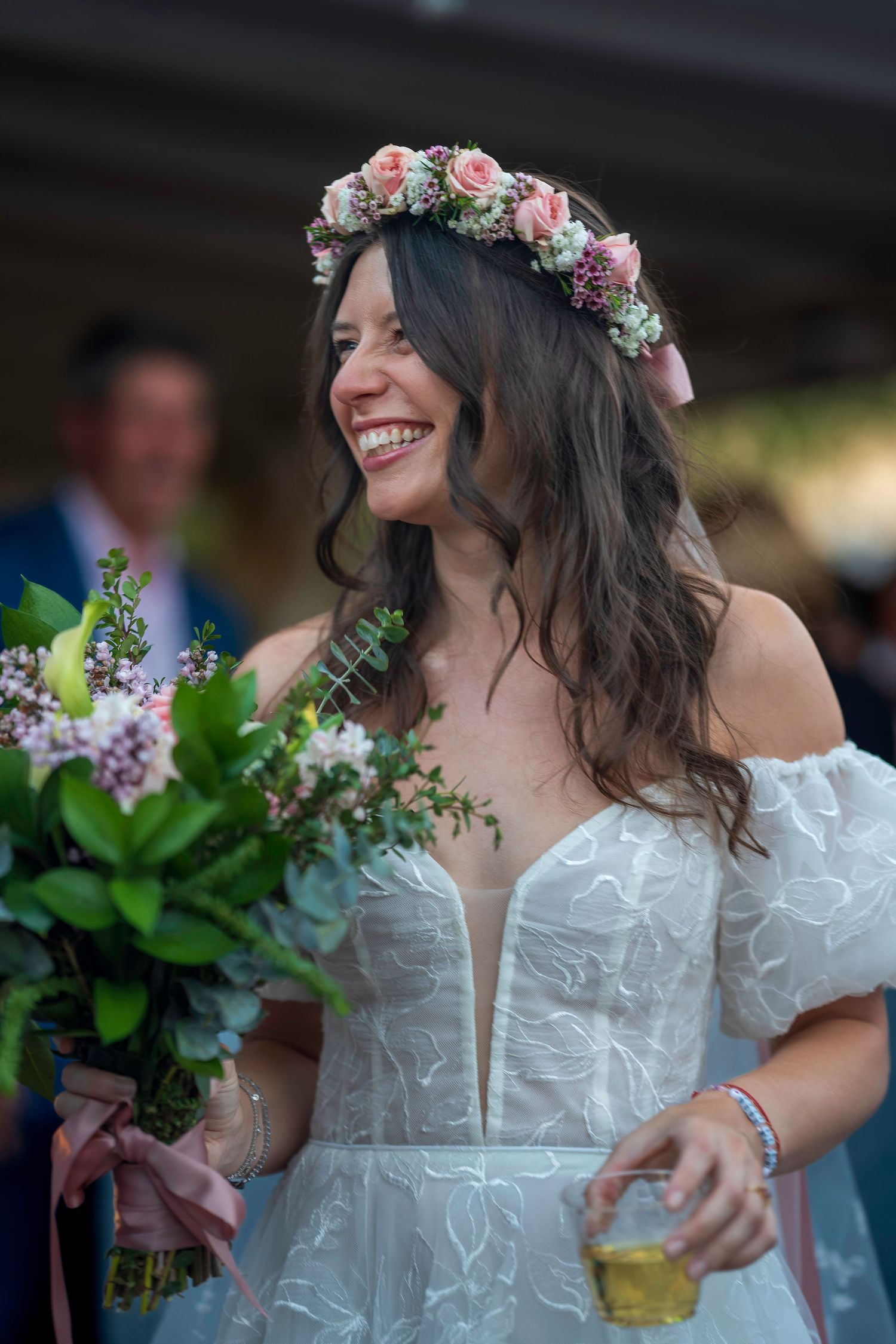 Smiling bride in white off-shoulder gown with floral crown holds bouquet at outdoor wedding ceremony.