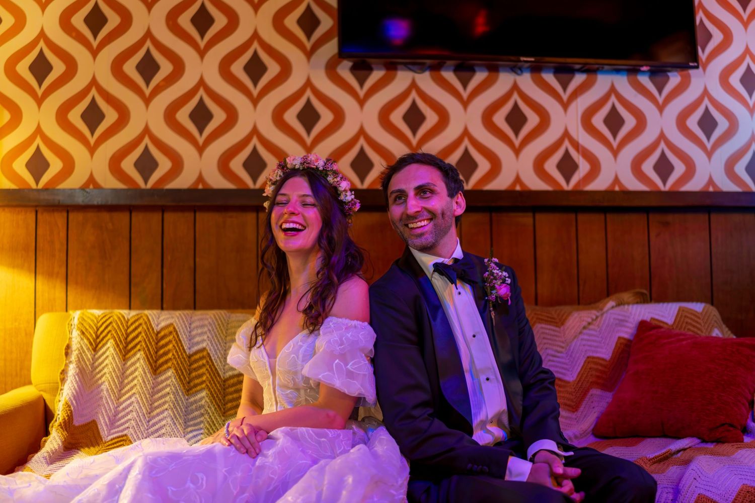 Bride and groom sitting together laughing on a bed with retro orange wallpaper backdrop at their wedding reception.