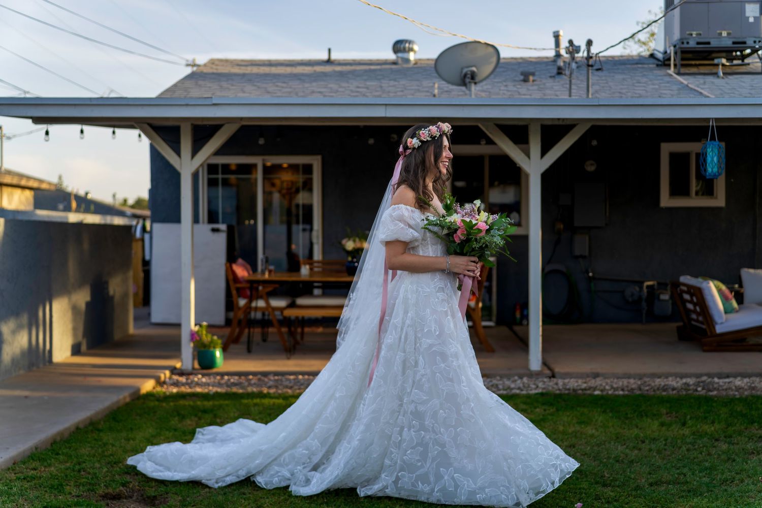 Bride in white lace gown holding floral bouquet stands in backyard before wedding ceremony.