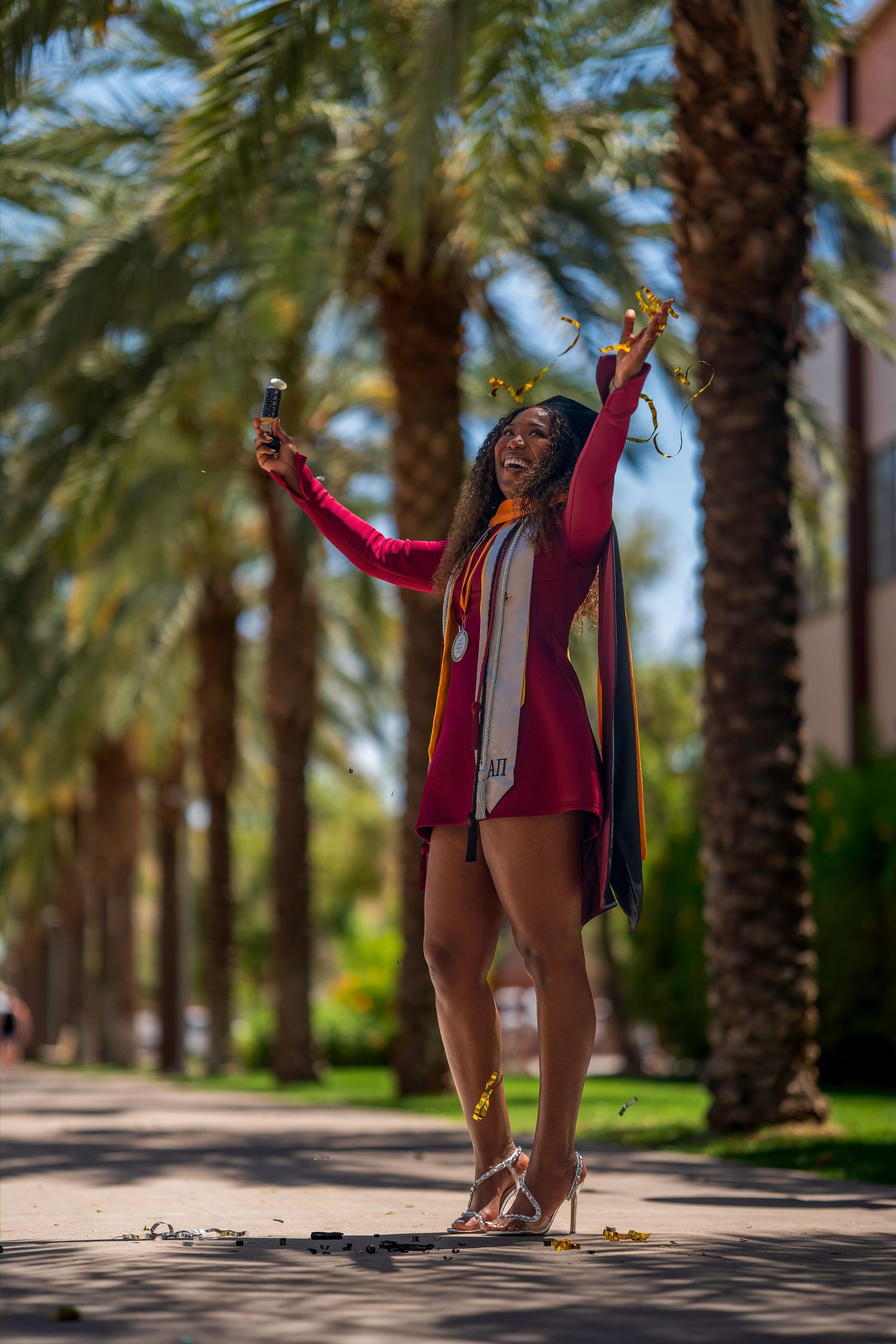 Joyful ASU graduate in maroon gown taking selfie along palm tree-lined campus walkway in heels.