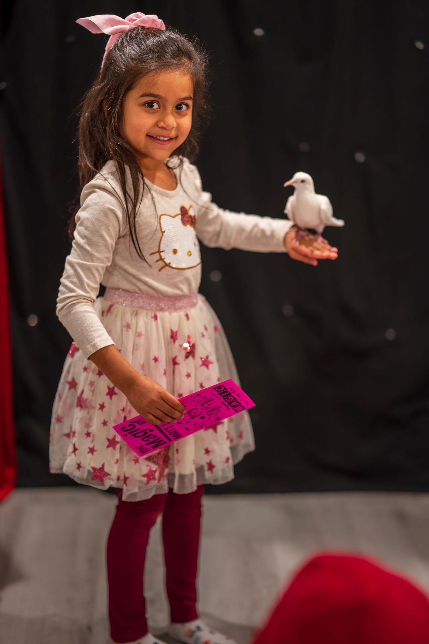 Child in a festive dress holds a white dove and pink prop against a dark backdrop.