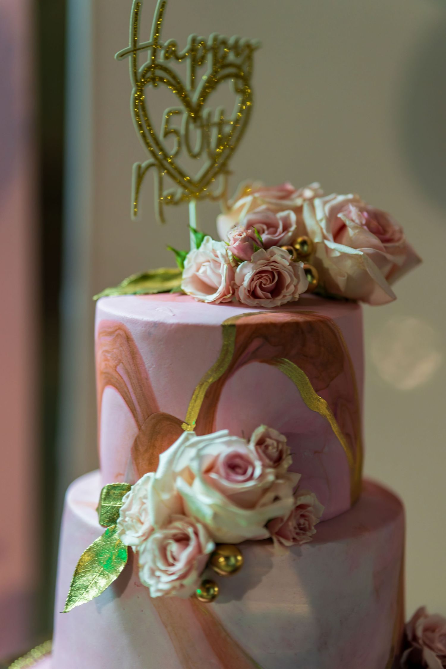 A pink wedding cake decorated with roses and gold 'Happy' topper details.