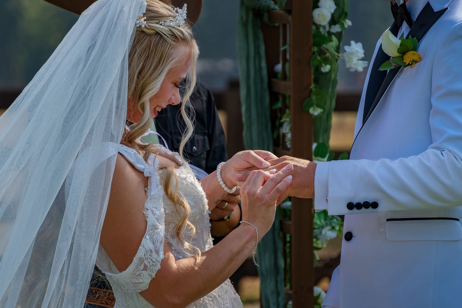 Wedding ceremony ring exchange moment with white floral arch and evergreen trees in background.